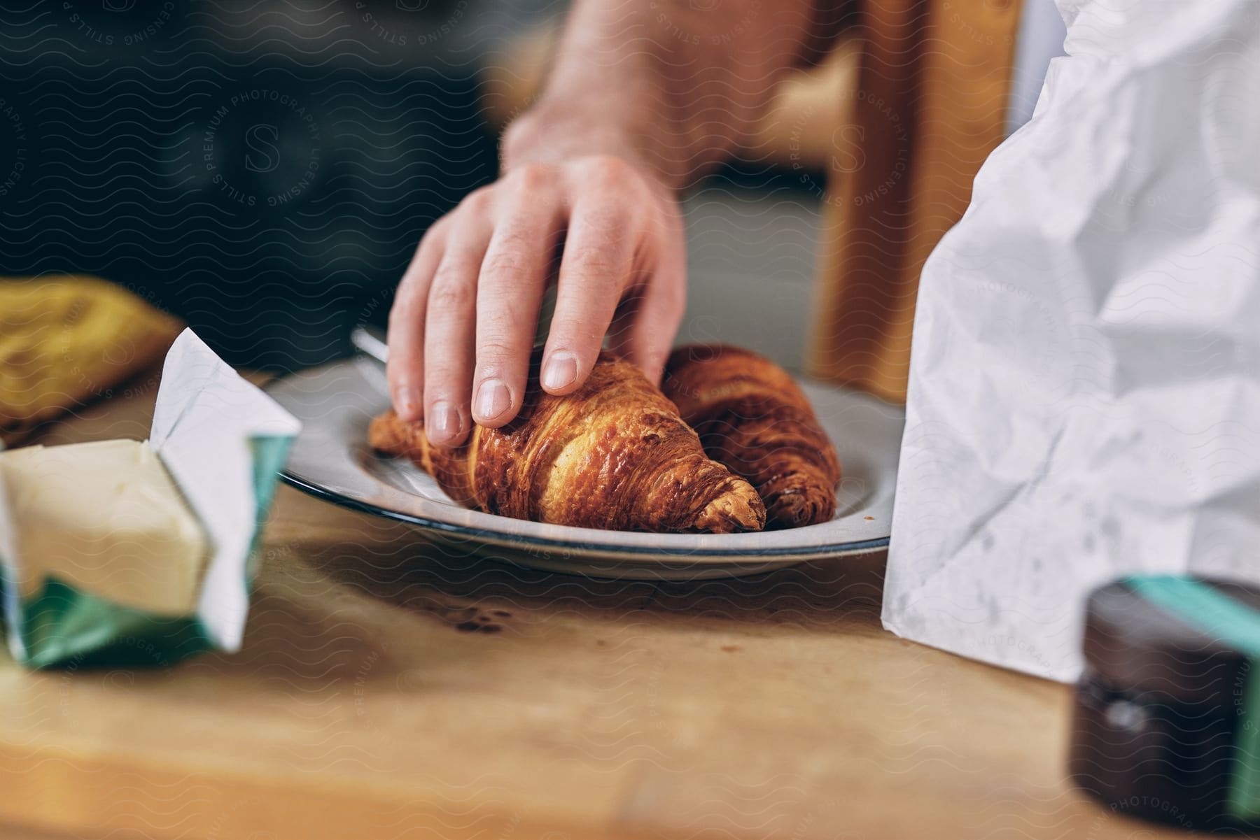 A person sets a croissant on a plate from a bag, next to another croissant, jam, and butter.