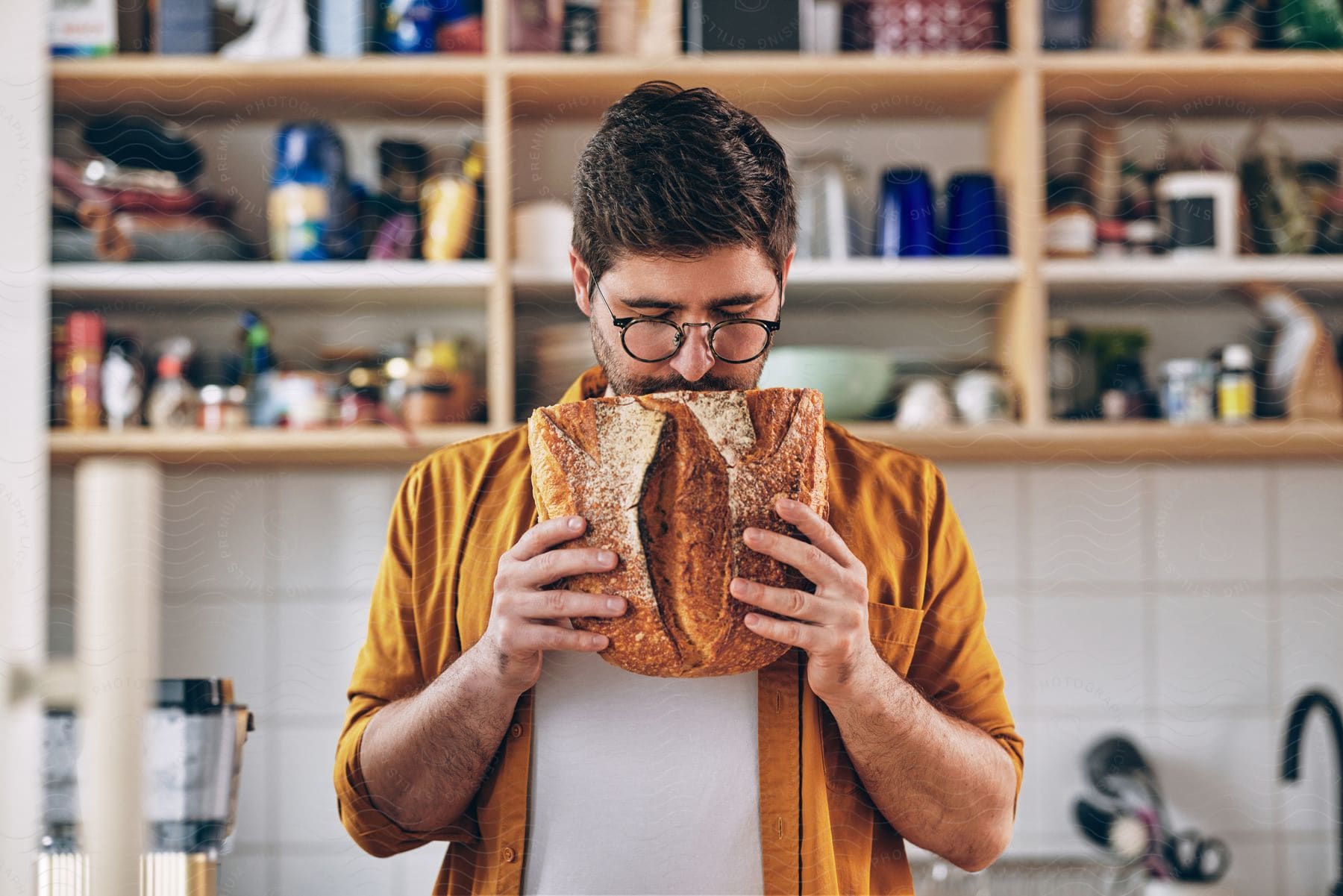 a man wearing a yellow shirt and a glass in the kitchen is sniffing a bread