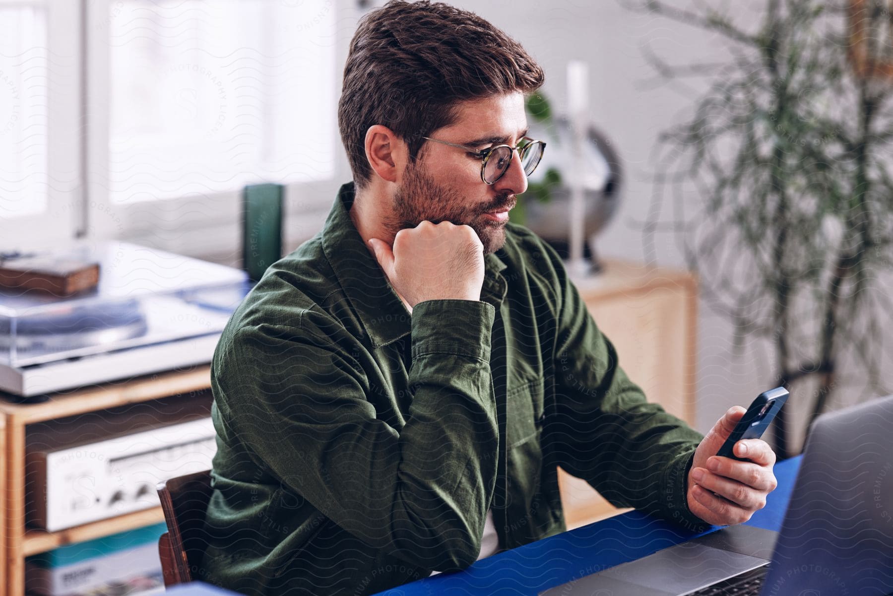 Stock photo of bearded man with glasses and a green shirt sits at desk using a smartphone and laptop in the office