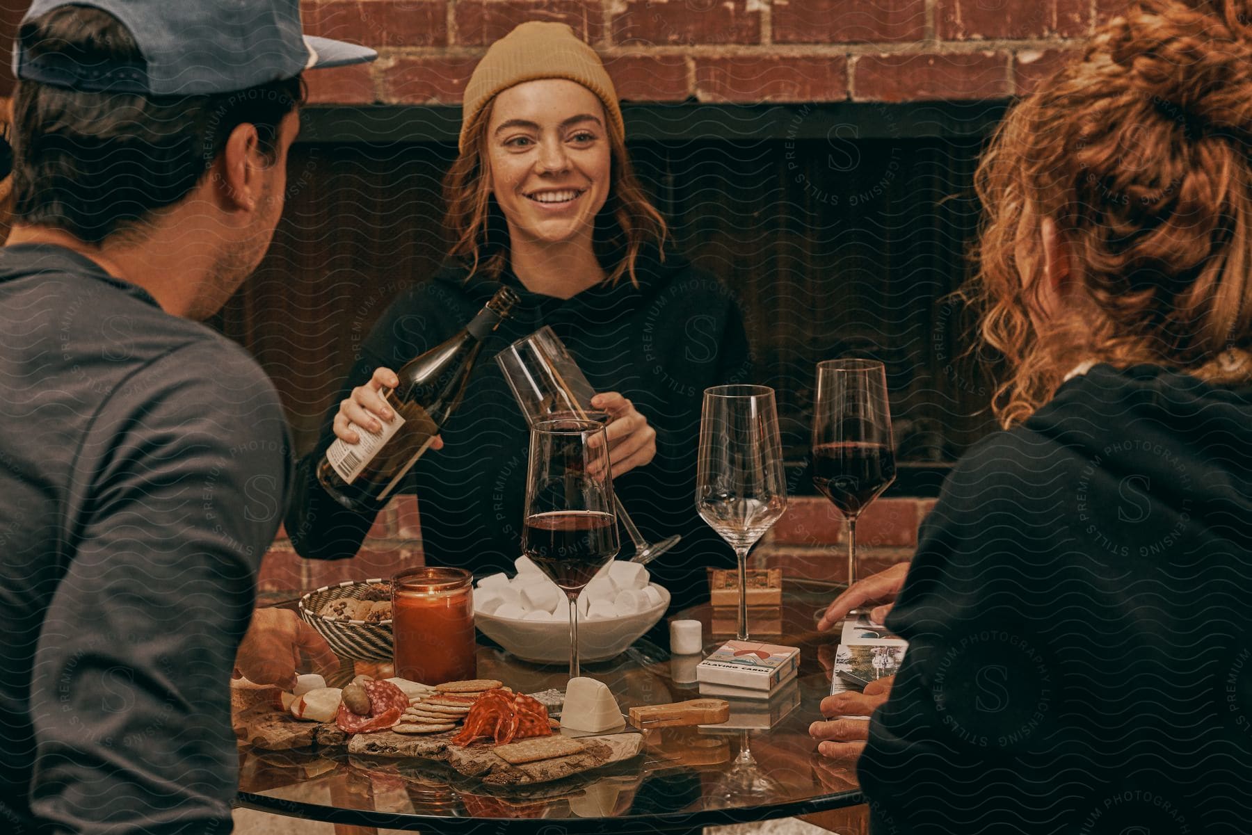 A woman holds a wine glass and a bottle of wine as she stands at a table smiling at two people