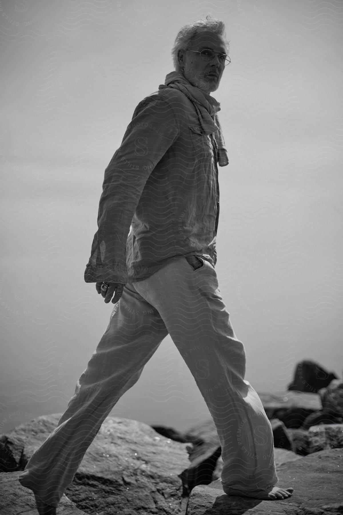 Man wears casual clothes while walking barefoot across rocks on shore.