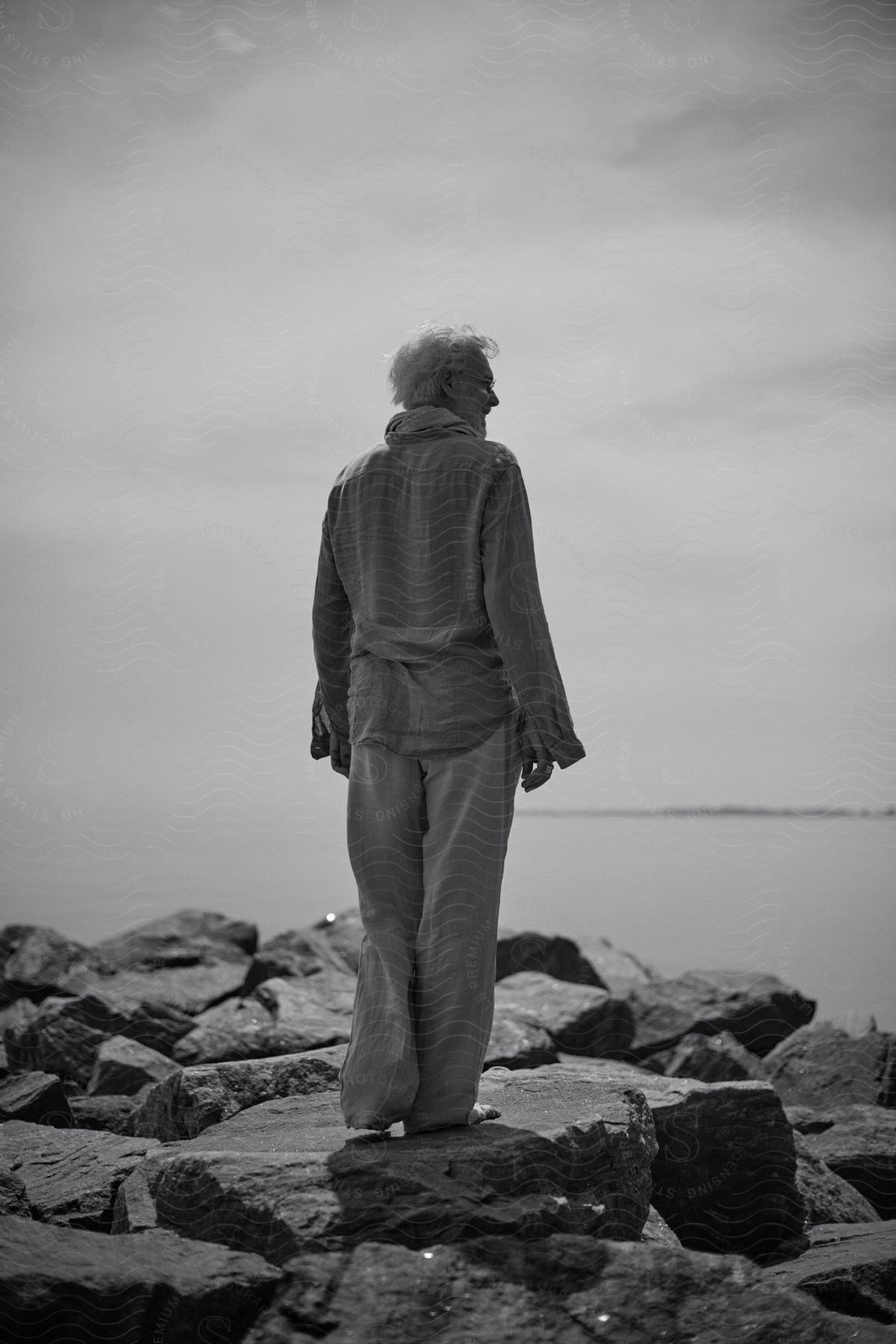 Older Man With Short White Hair Stands On The Rocks Next To The Ocean