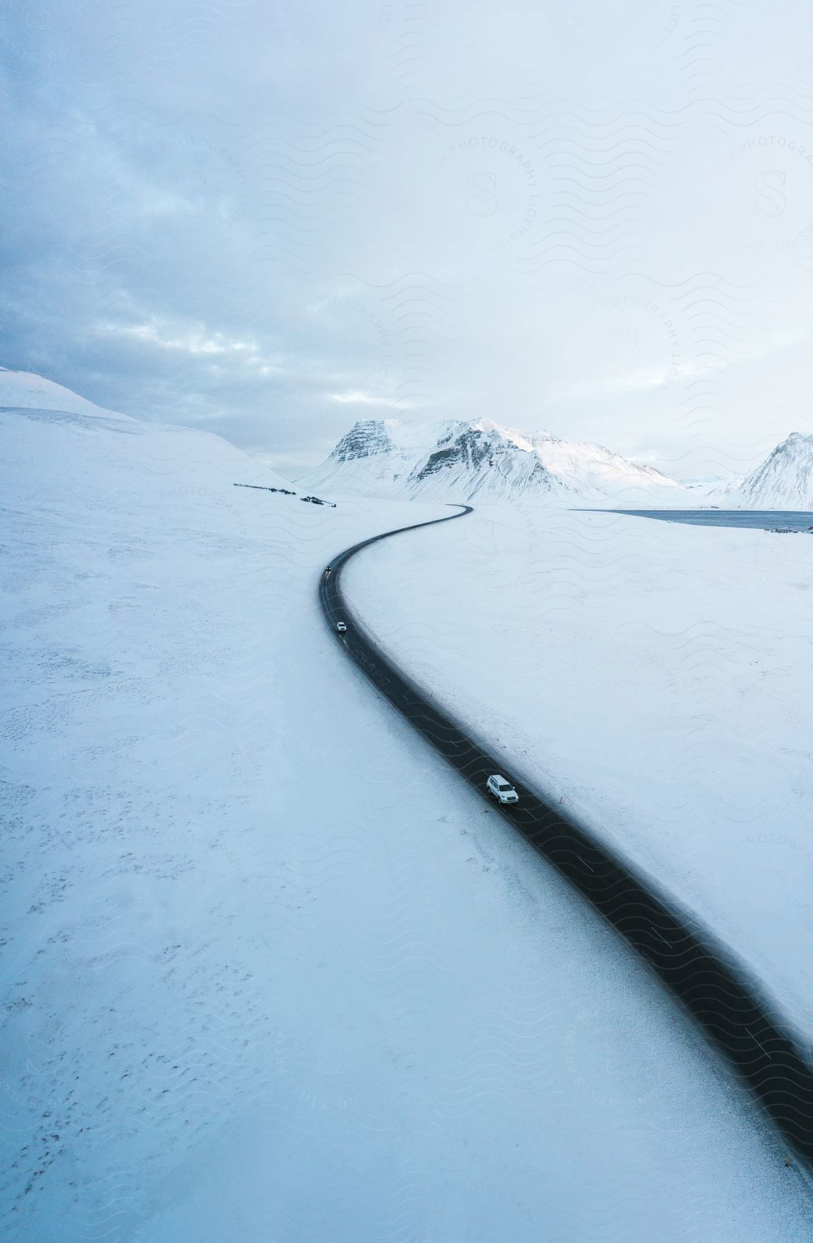 A white SUV snakes through a freshly plowed mountain pass, snow-covered peaks rising in the distance with snow covering the ground.