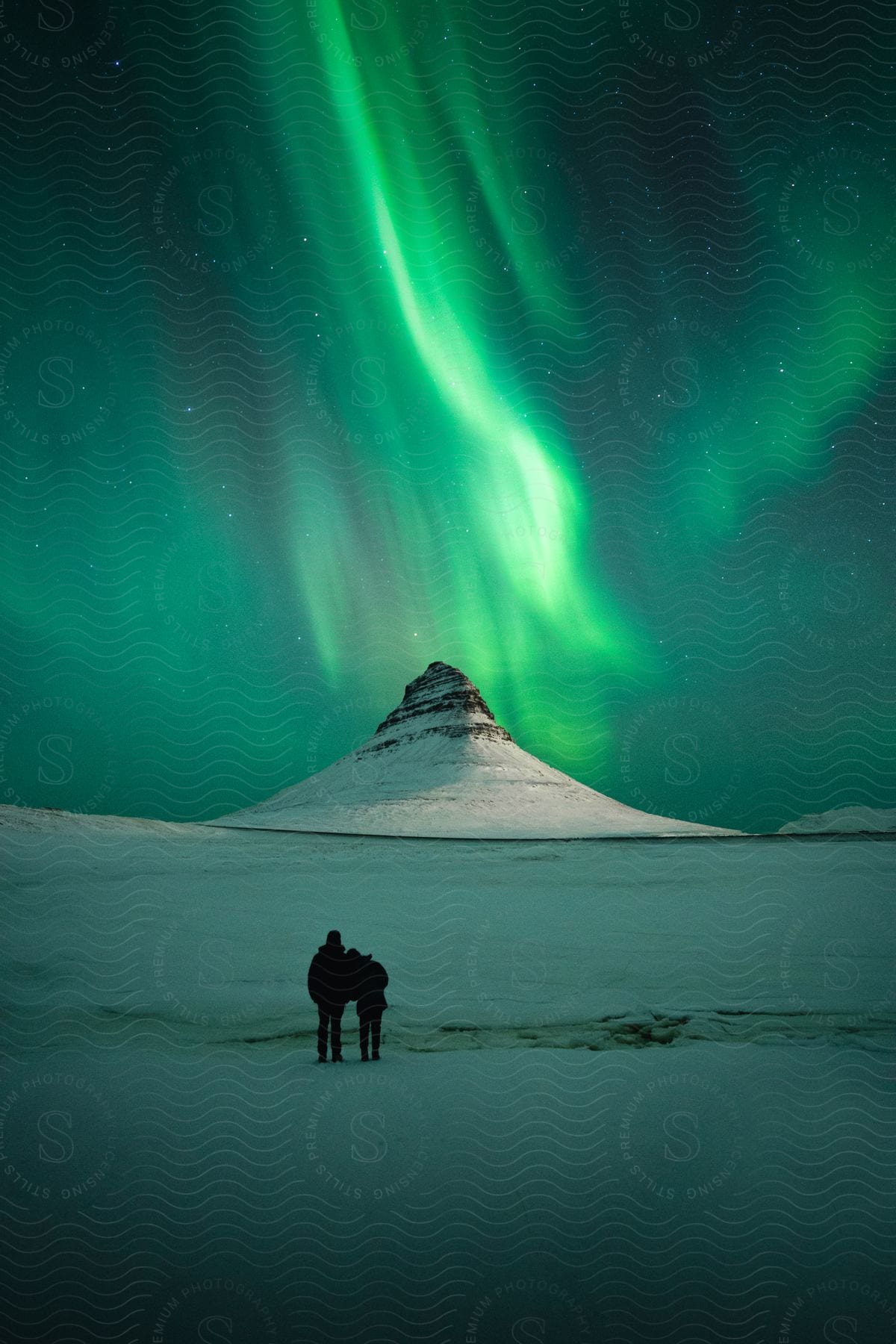 A Couple Lean Against Each Other While Watching The Aurora Borealis Over A Mountain Peak