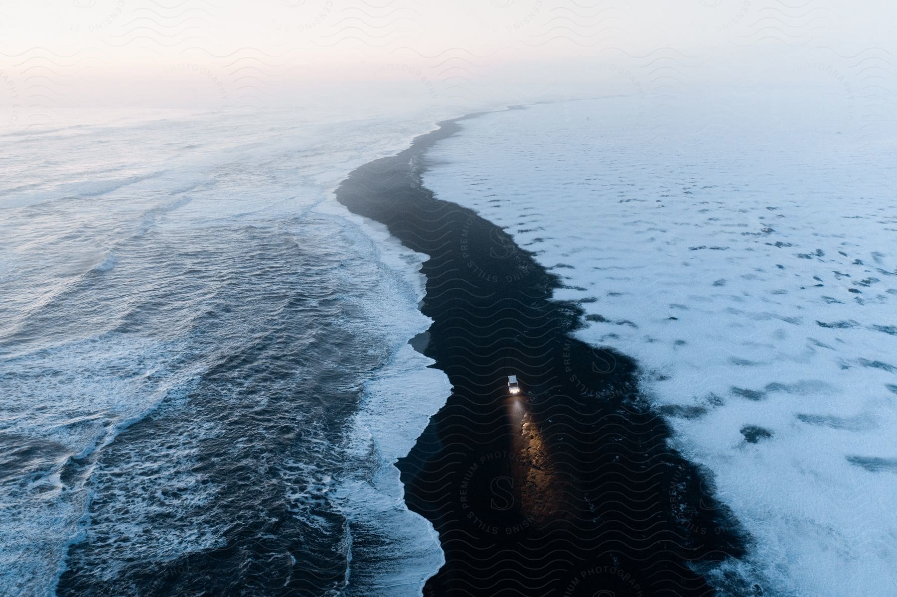 An aerial view of a white car driving along a black beach near the ocean waves.