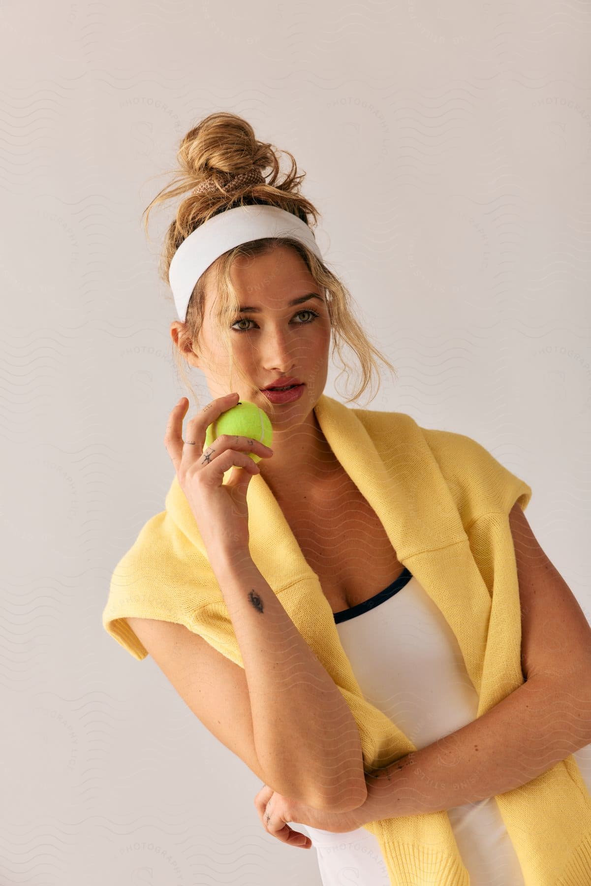 A woman in white athletic wear with a yellow sweater draped over her shoulders holds a tennis ball, posing against a neutral background. She's wearing a white headband and her hair is styled in a bun.