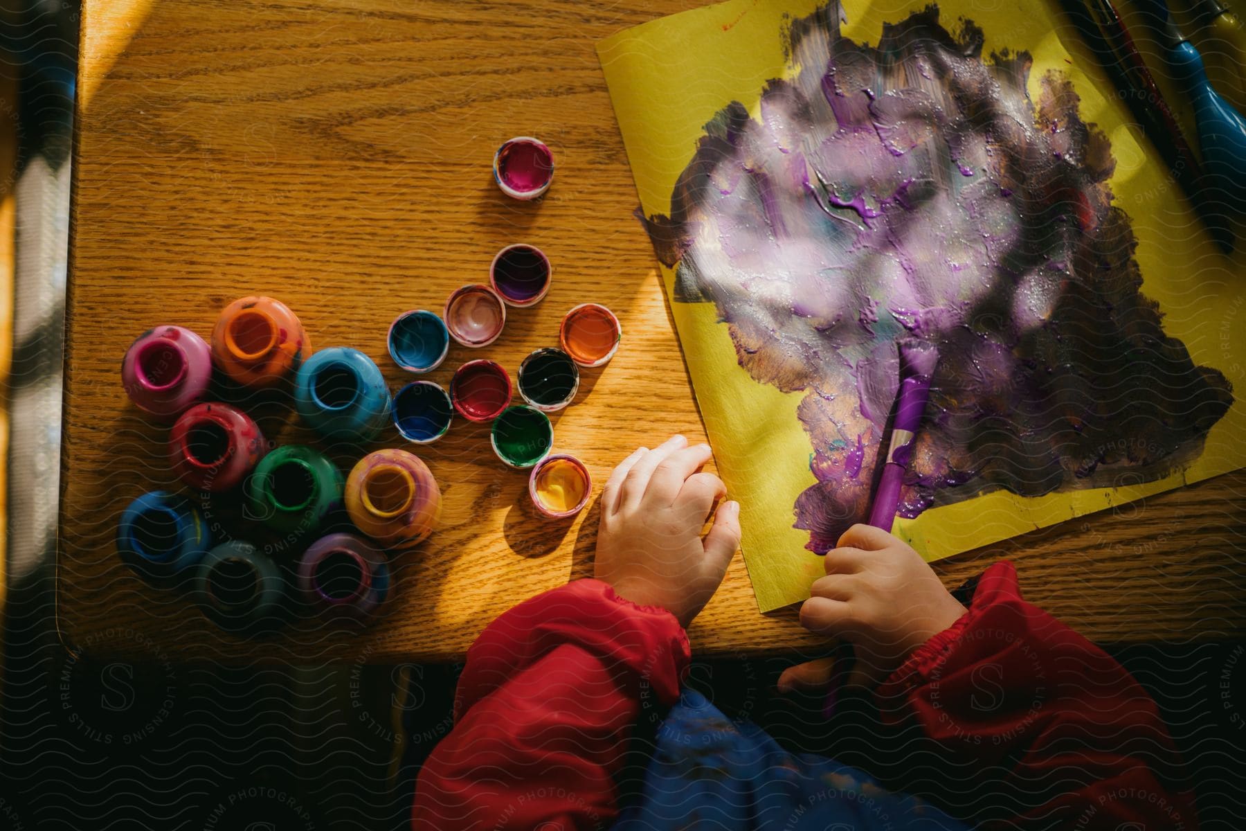 A child is holding a paint brush and painting a picture with a hand on the table near many colored paint containers