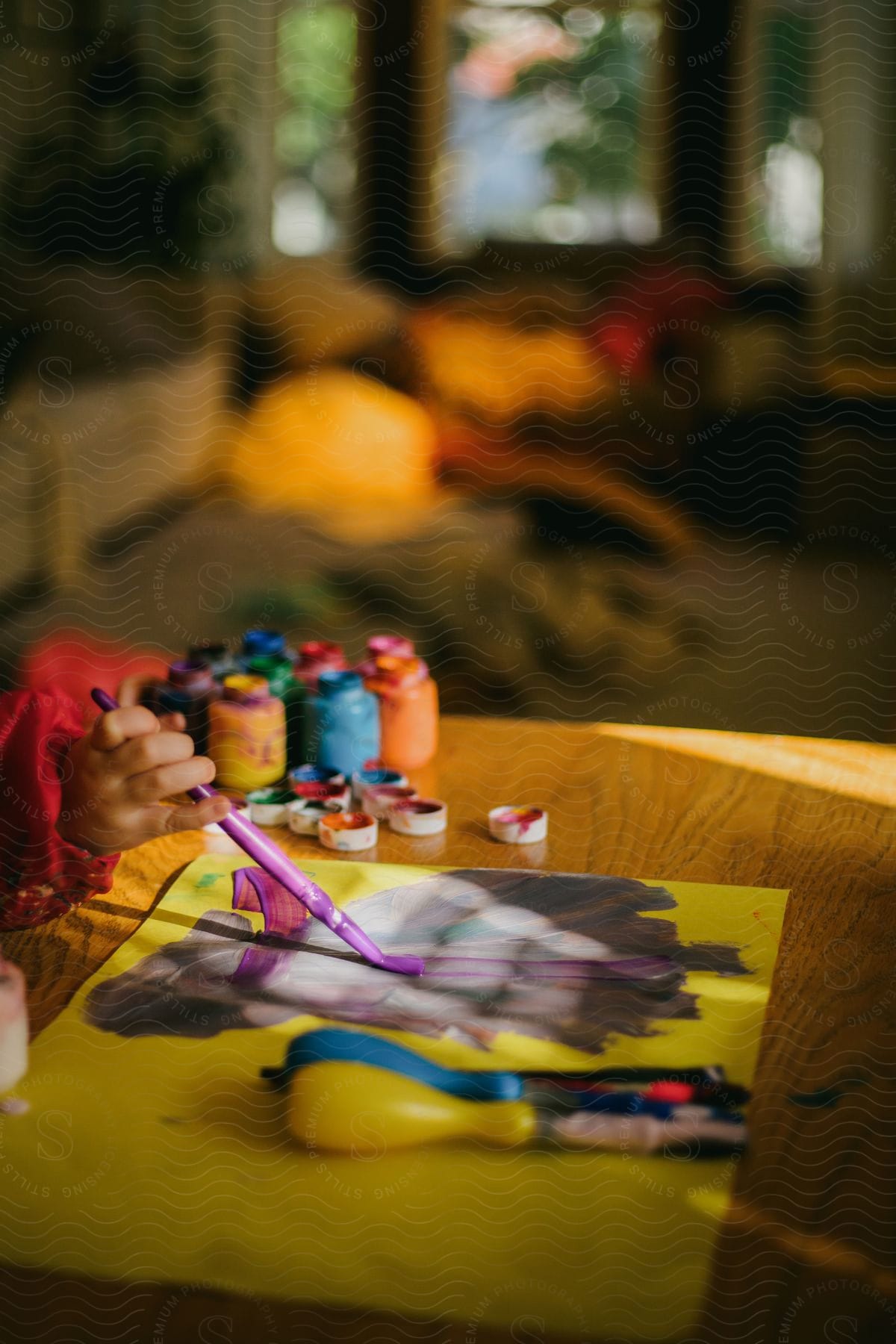 A child is holding a paint brush and painting a picture with many colored paint containers on the table