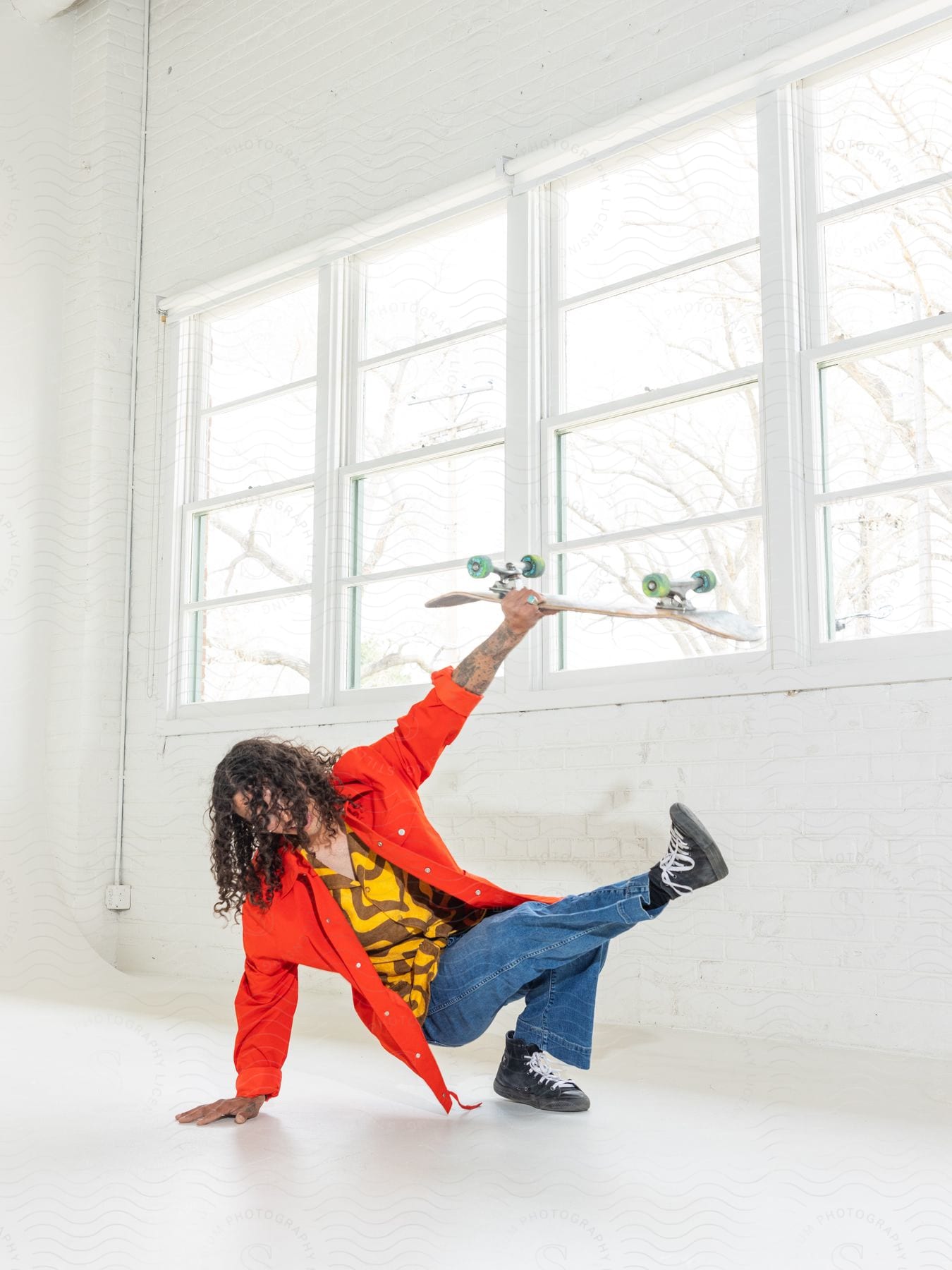 A man has one hand on the floor and a foot in the air as he holds up his skateboard in a white room