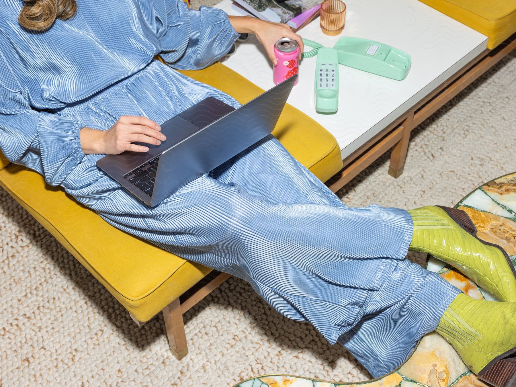 A woman is sitting in a chair typing on a laptop computer sitting on her legs as she holds a can of pop near a phone on the table