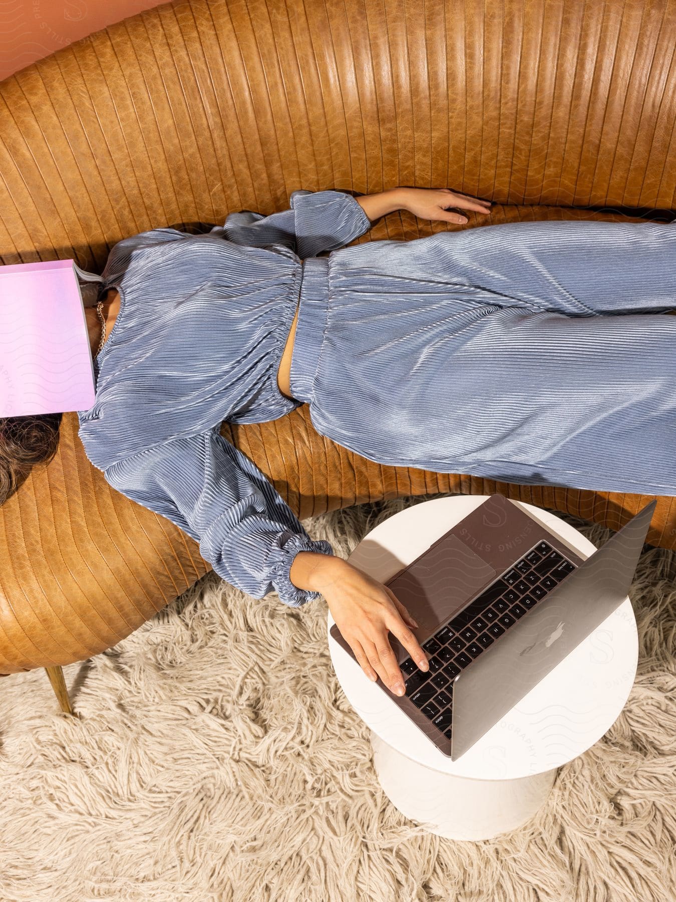 A woman in a corduroy sweat suit lying on a couch with a book over her face and her hand on a laptop.