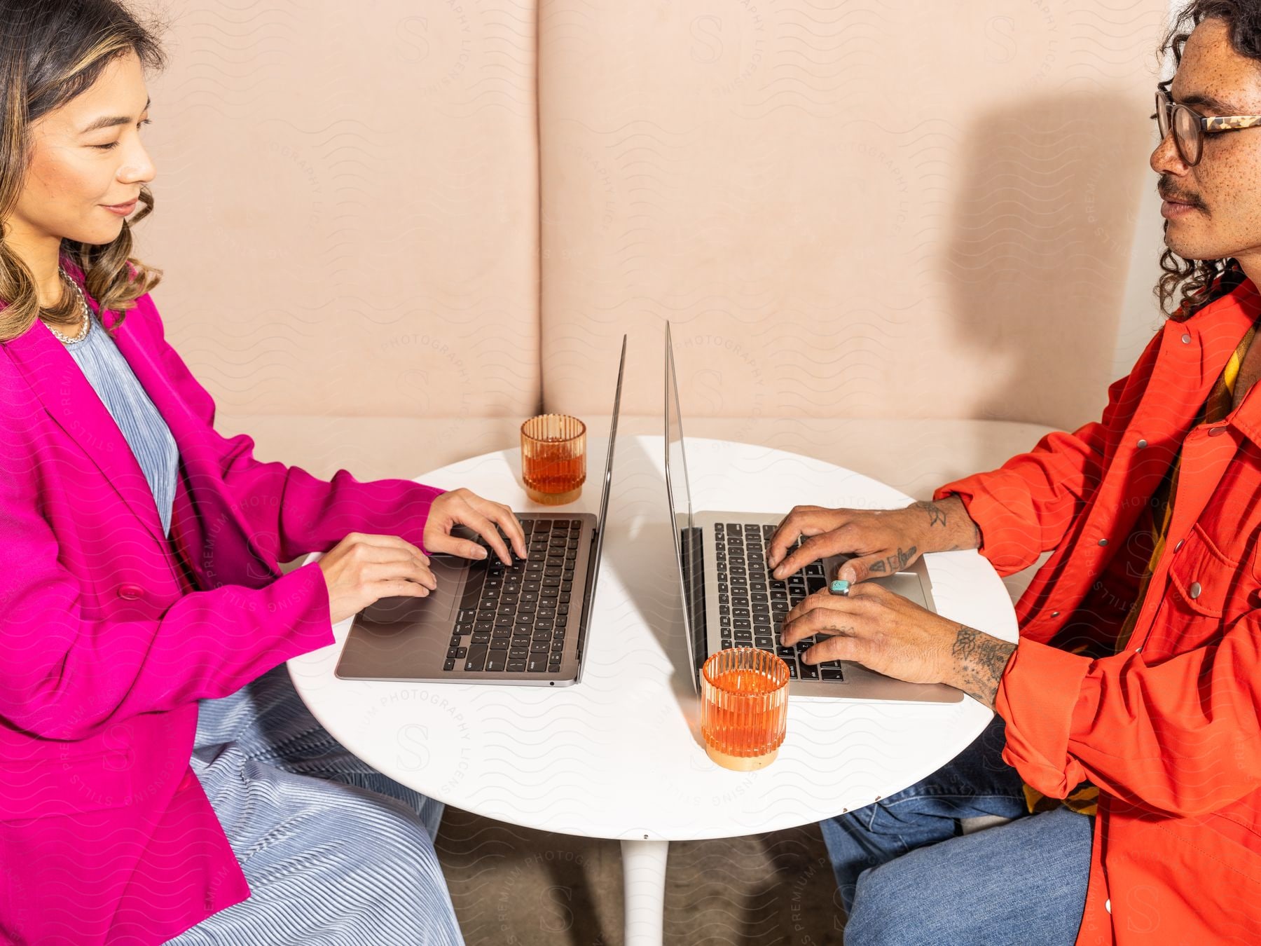 A man and a woman sitting at a table typing on laptops.