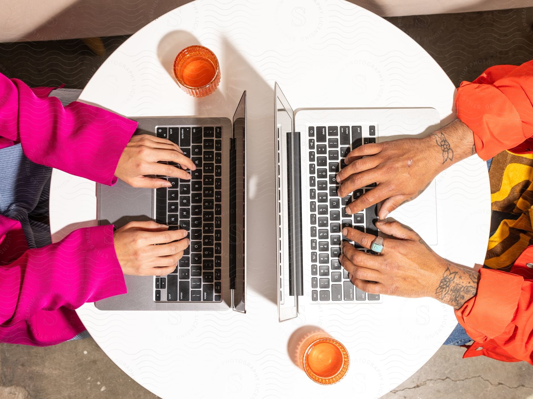 Two people are sitting at a round white table facing each other as they type on laptop computers with a cup on each side