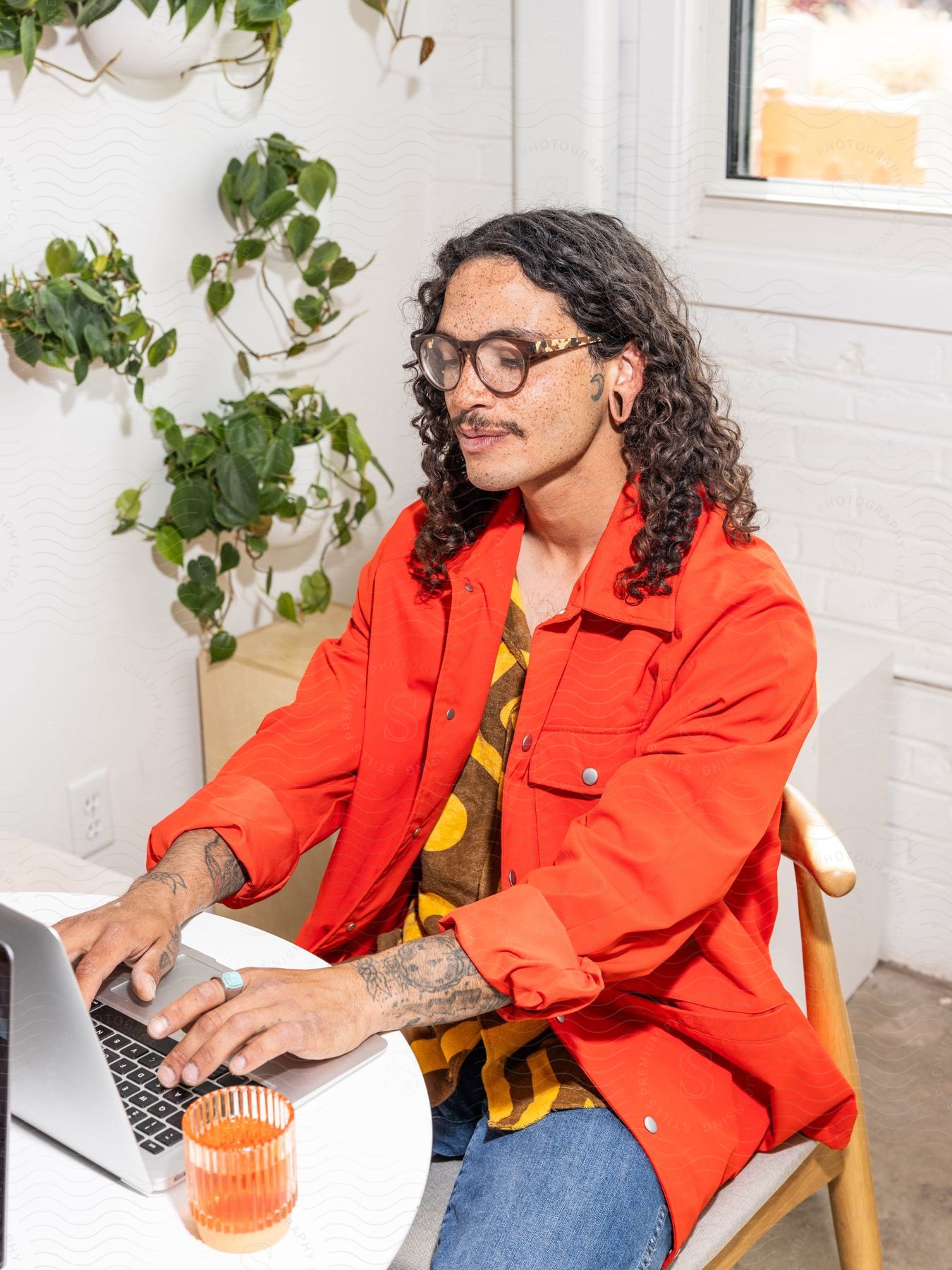 Man in red jacket and glasses working on a laptop at a white table with a cup beside him, against a white wall with hanging green plants.