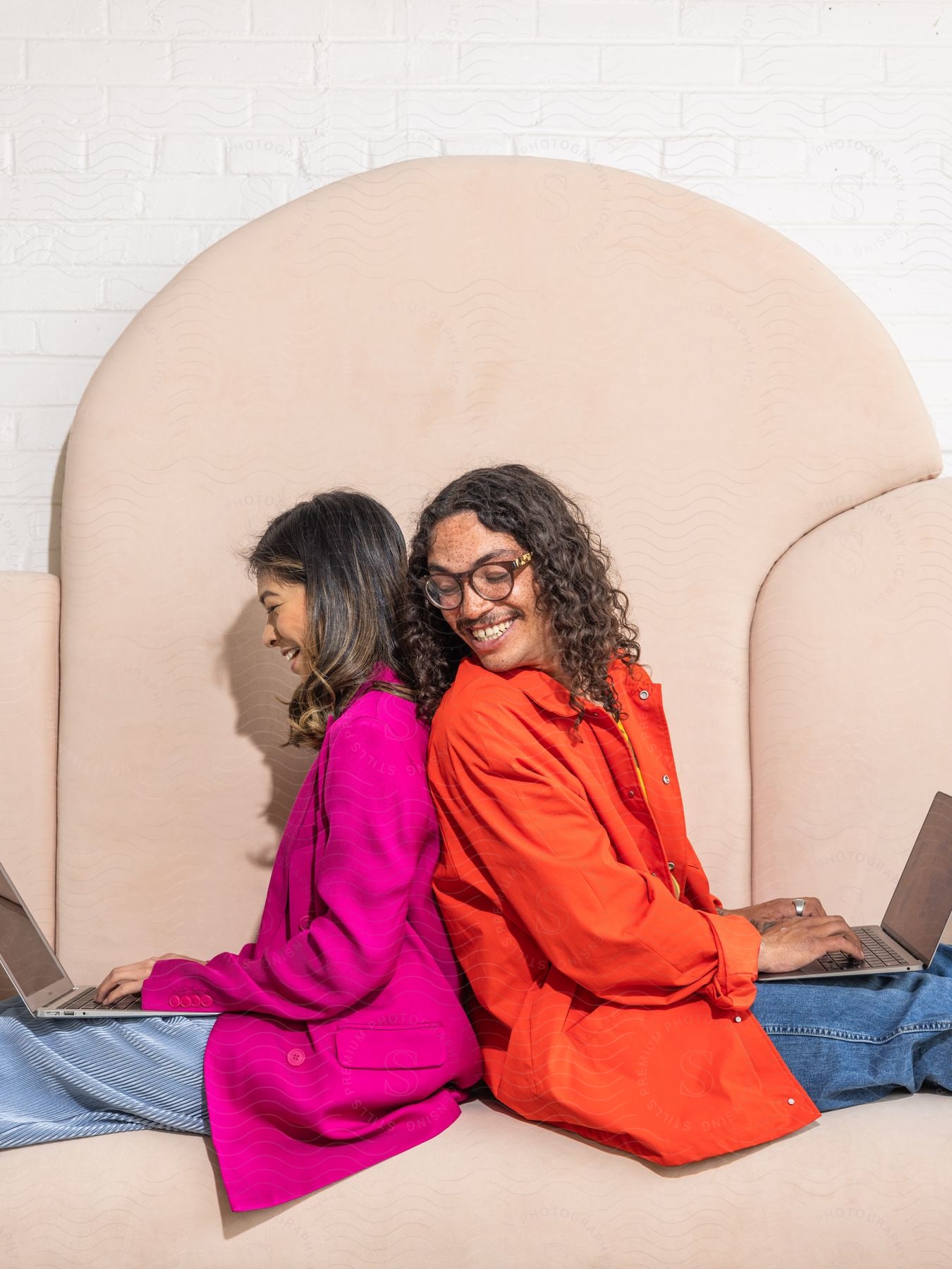 Stock photo of a young man and woman are sitting back-to-back and smiling while holding laptops on their laps.