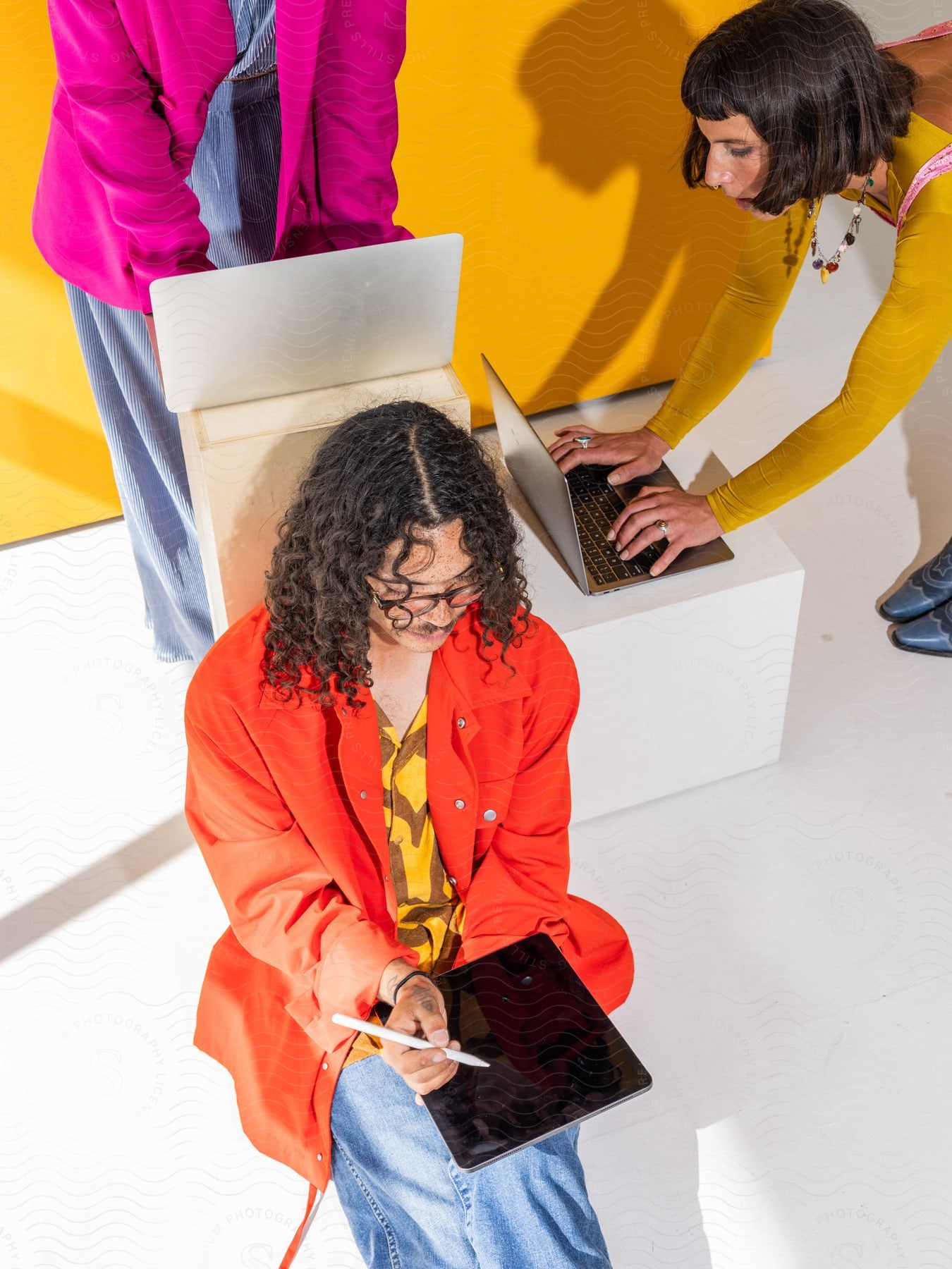 Three adults are using laptops and a tablet while wearing bright-colored clothing.