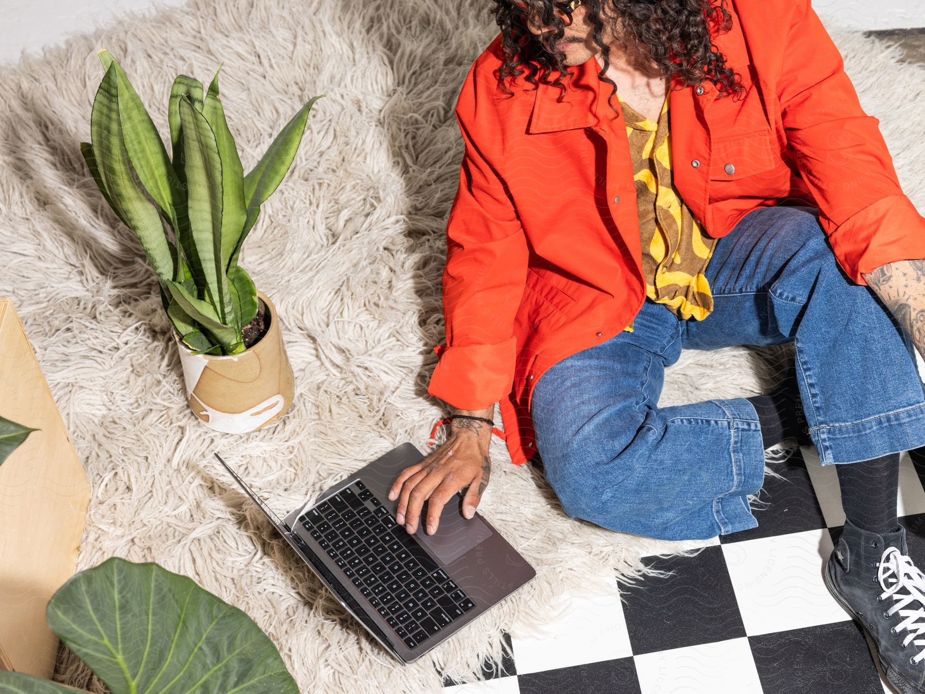 A man sits on a rug with potted plants and his hand on a laptop computer keyboard