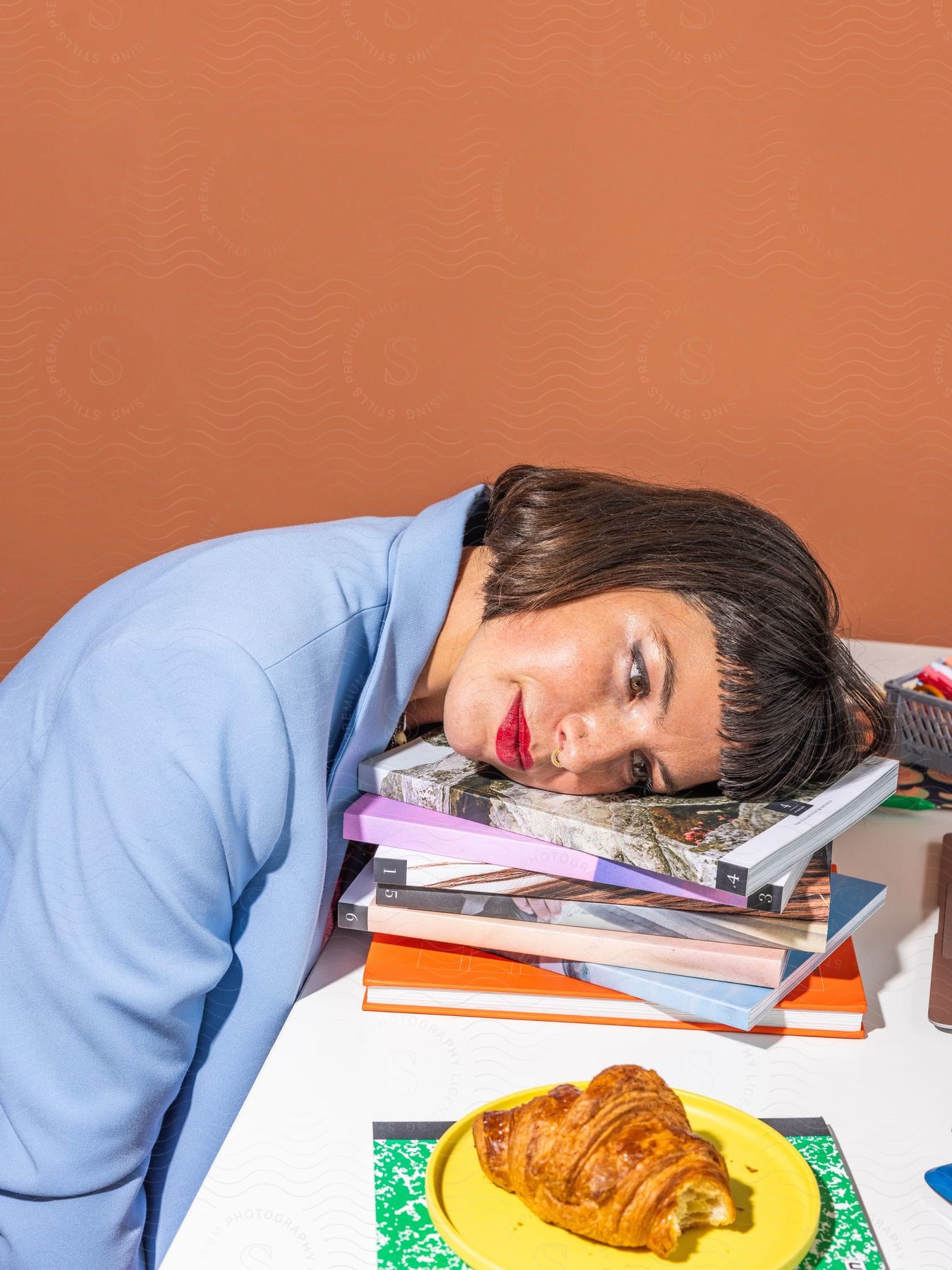 A woman sitting at a desk is resting her head on books looking at a pastry on a plate