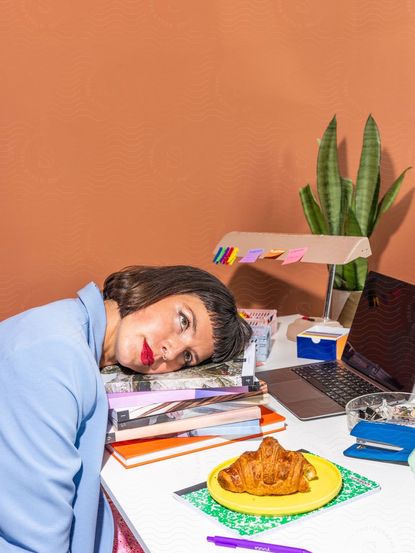 Woman resting her head on a stack of books at a desk, with a laptop, plant, and croissant on a plate nearby, against a peach-colored backdrop.