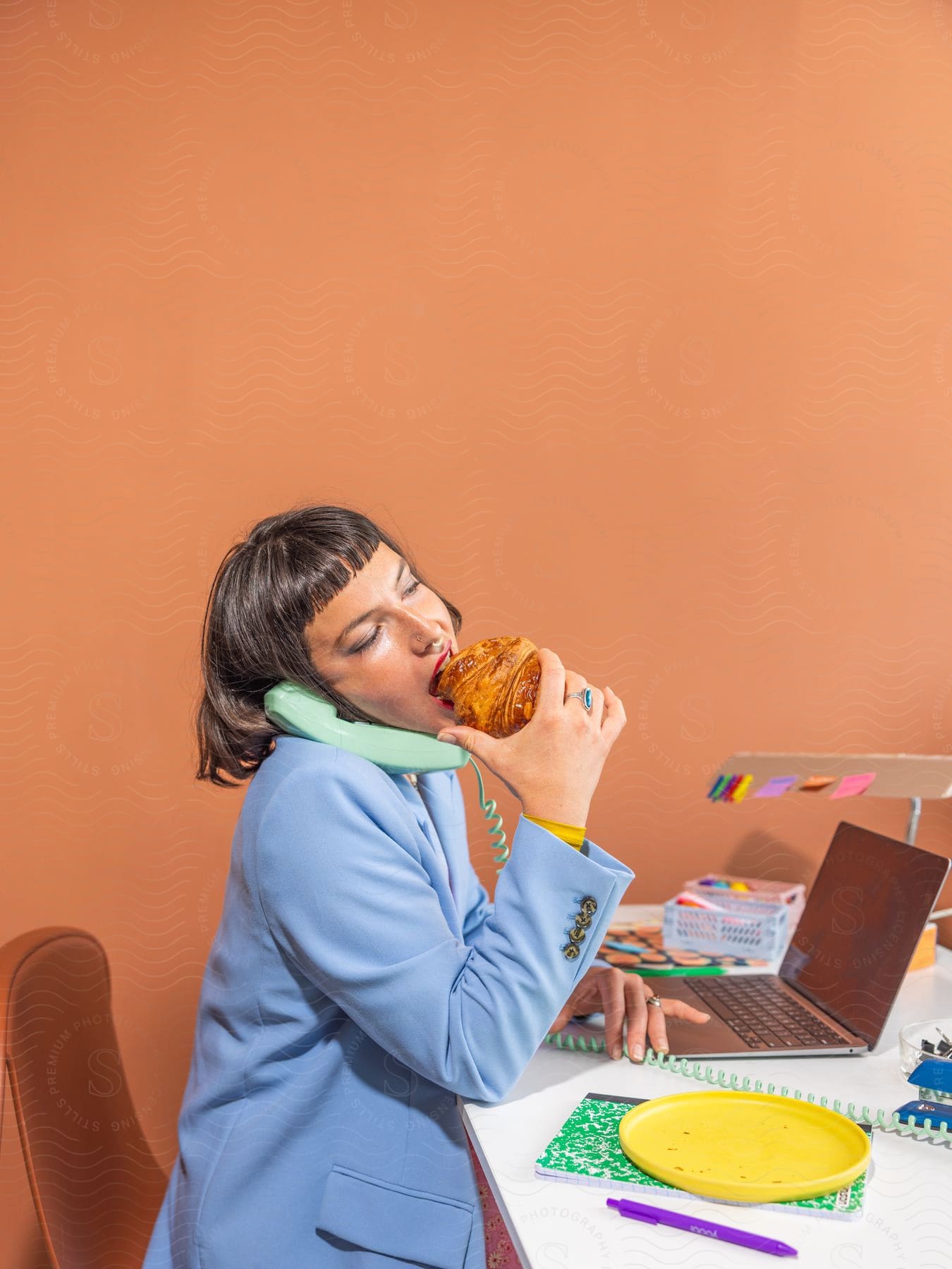 Modeling woman wearing a light blue blazer and pretending to eat a croissant while answering a phone call.