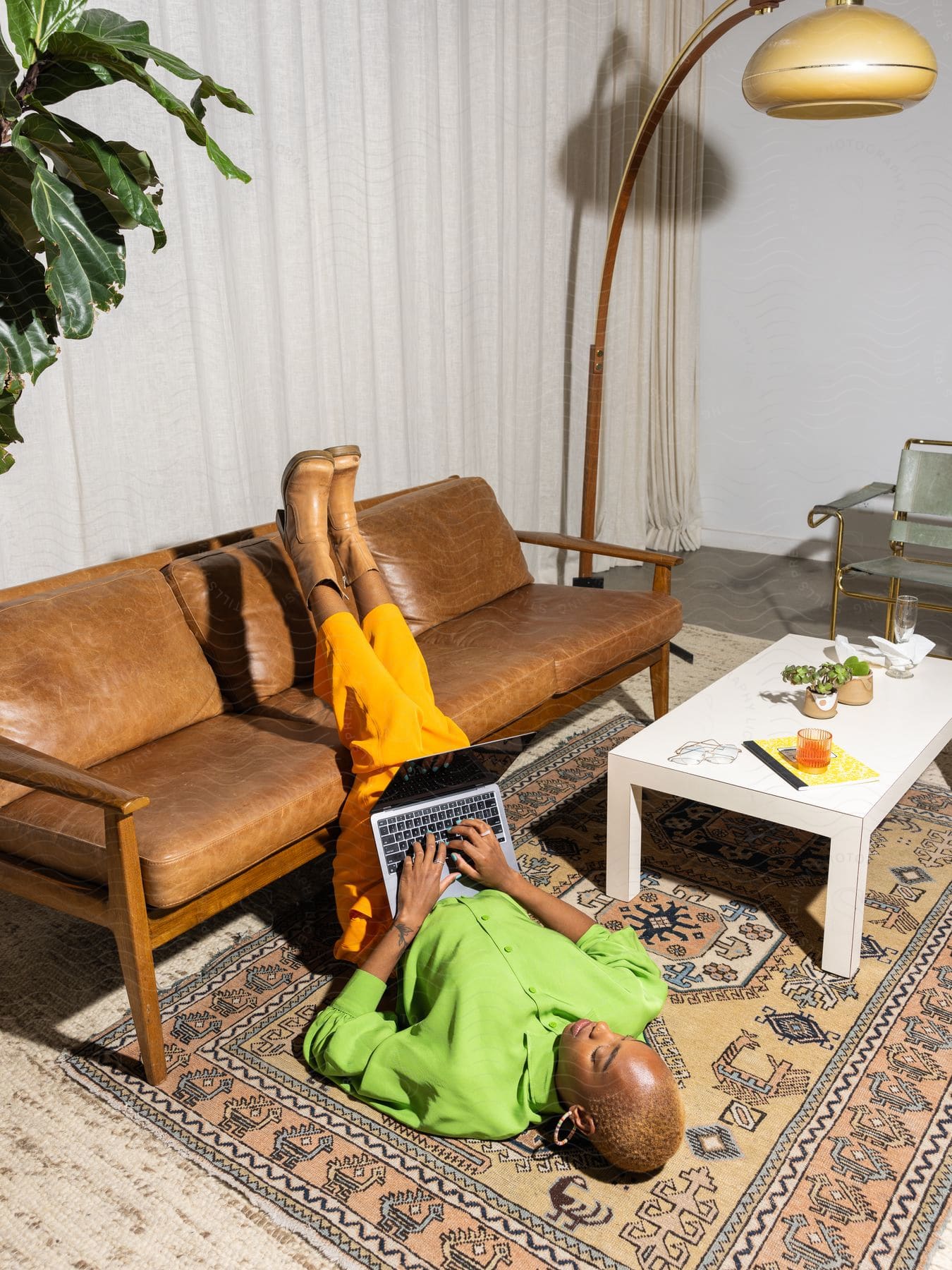 Woman in green top and yellow pants lying on her back on a rug, legs on a brown sofa, using a laptop, in a room with a floor lamp and a coffee table with items.