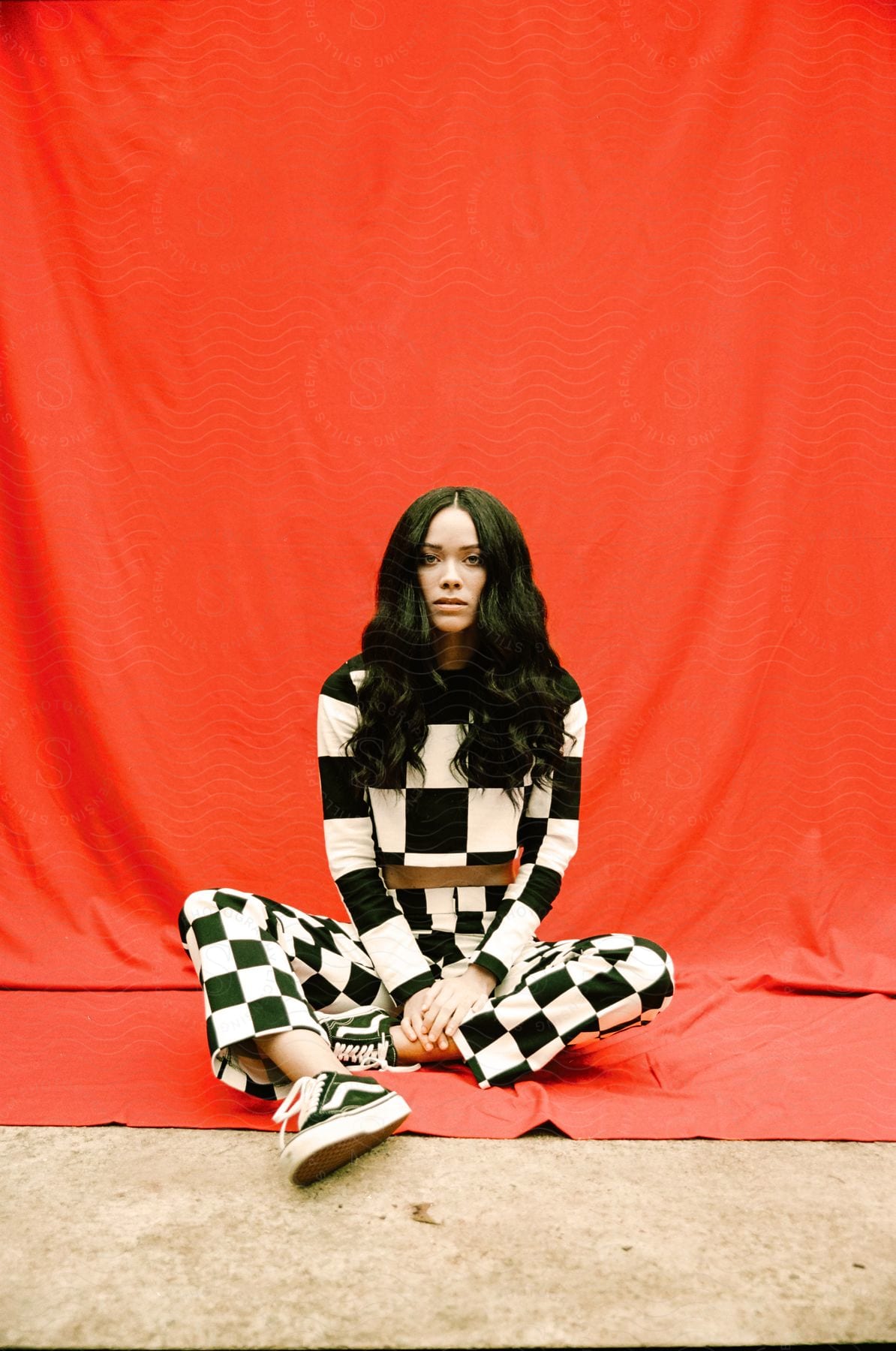 Woman wearing checked patterned outfit sits at base of red canvas backdrop in photography studio.