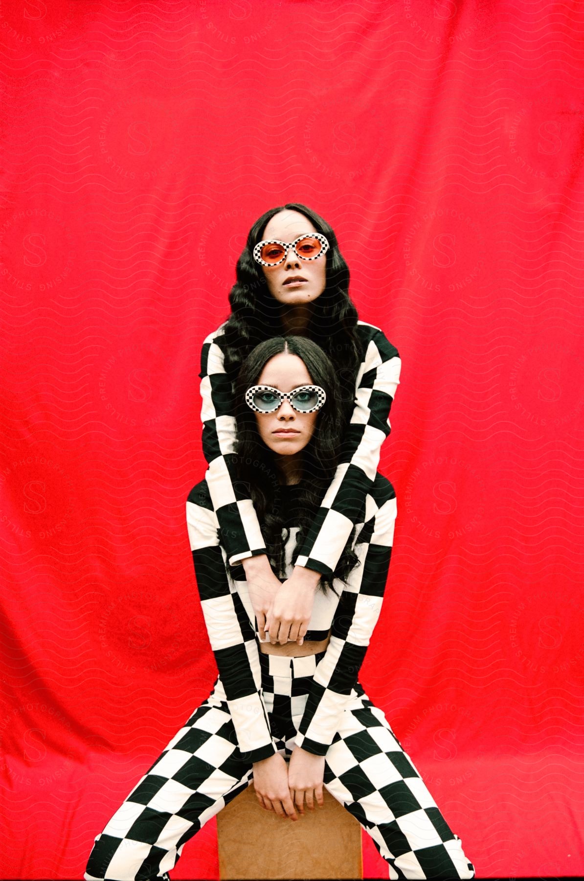 two young ladies wearing the same black and white outfit with one sitting and the other behind her with a red background behind them