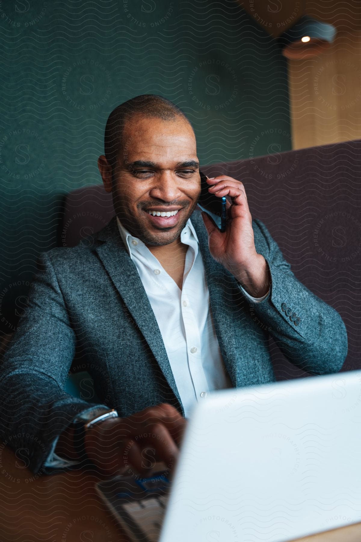 A man wearing a blazer smiles as he is on the phone while using his laptop computer.