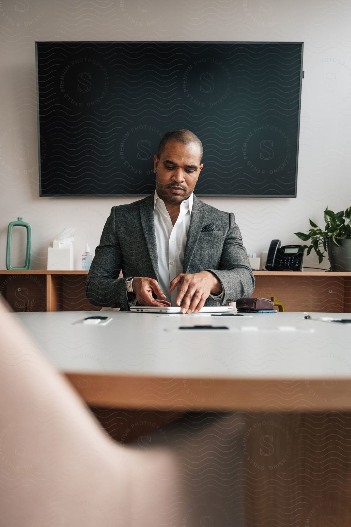 a black man in suit, seated in a conference room with a television at his back