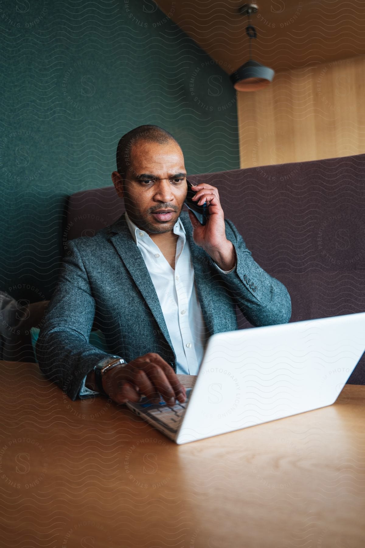 A man sits and focuses on his laptop computer while he holds his smart phone to his ear.