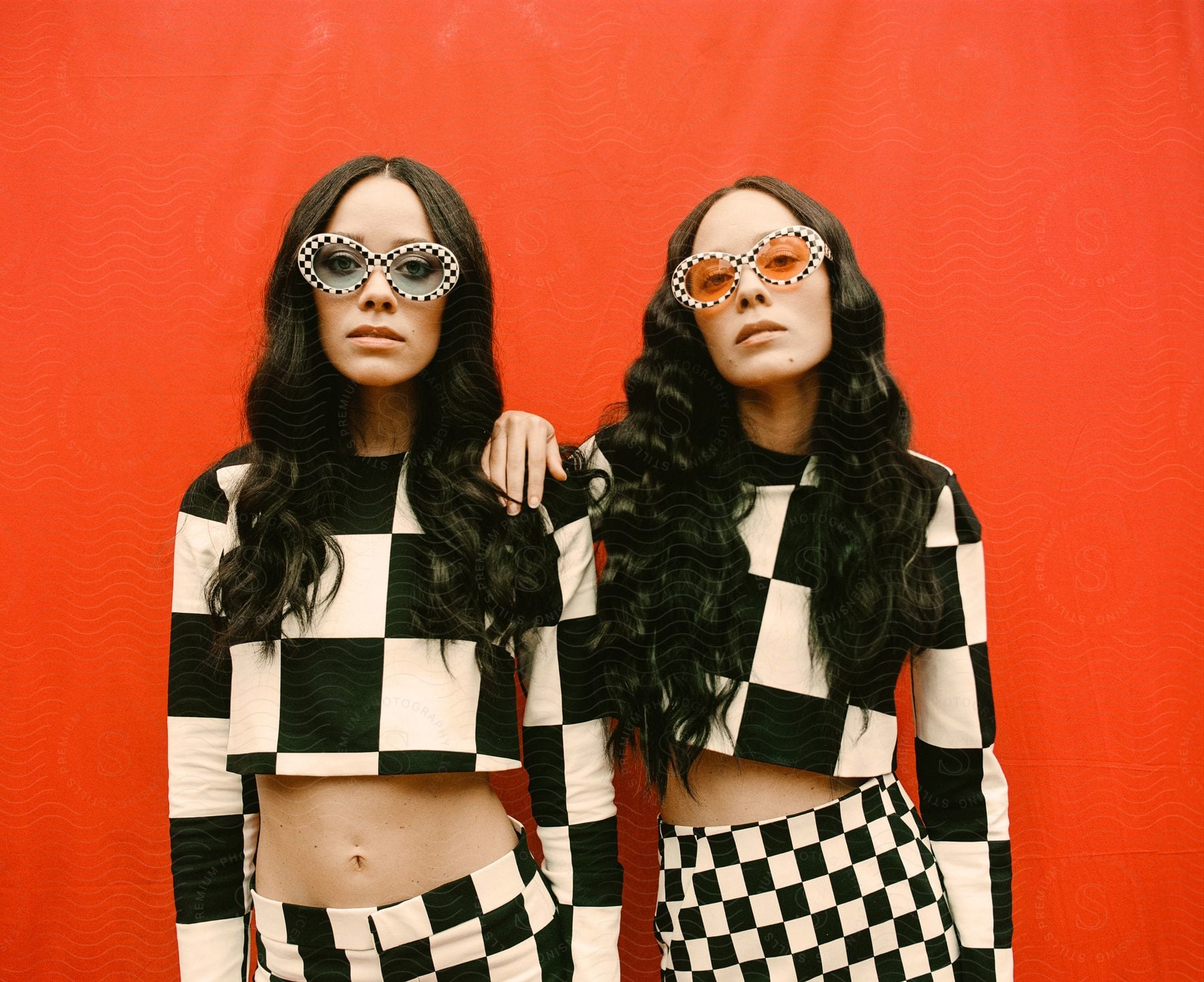 Portrait of two women posing in identical outfits against a red background.