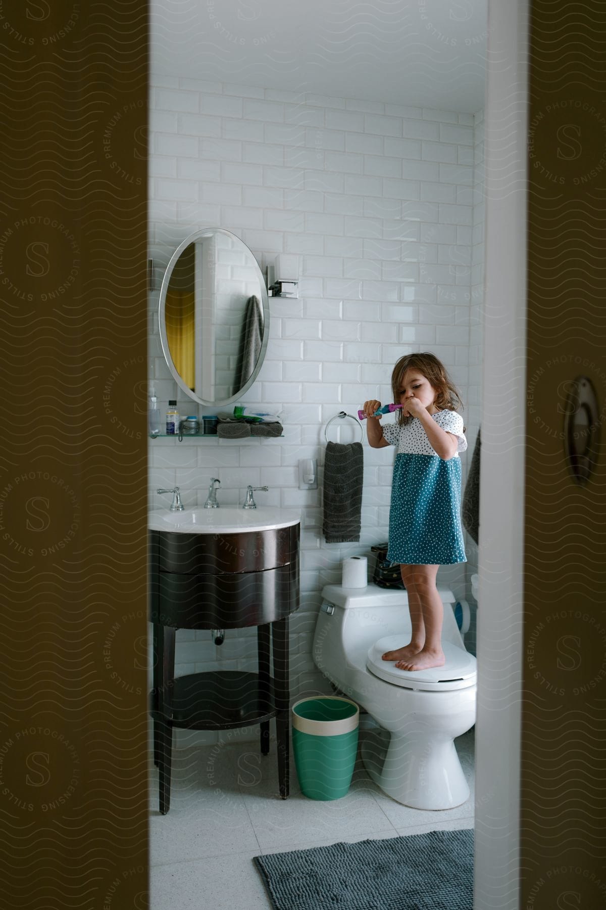 A little girl wearing a dress is standing on the toilet near the bathroom mirror