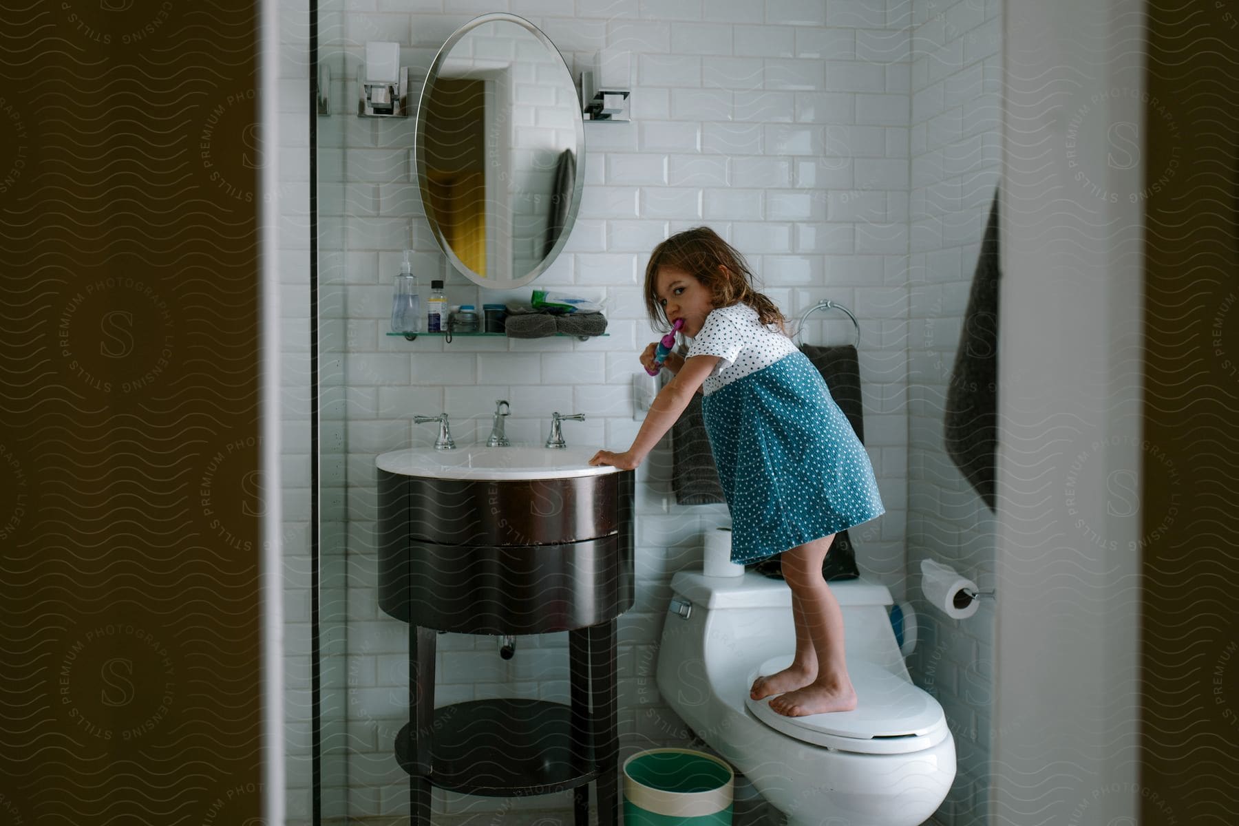 A young girl standing on a toilet so she can wash her teeth in the bathroom sink.