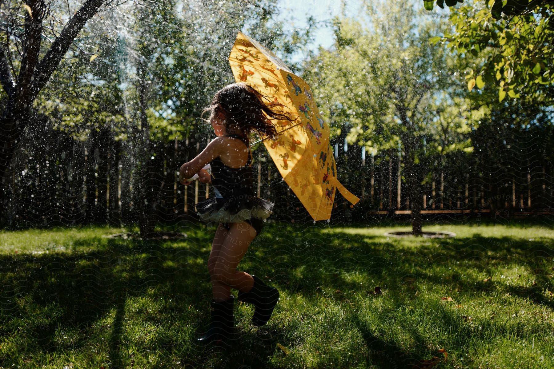 A little girl is holding an umbrella as she plays in a water sprinkler in her yard