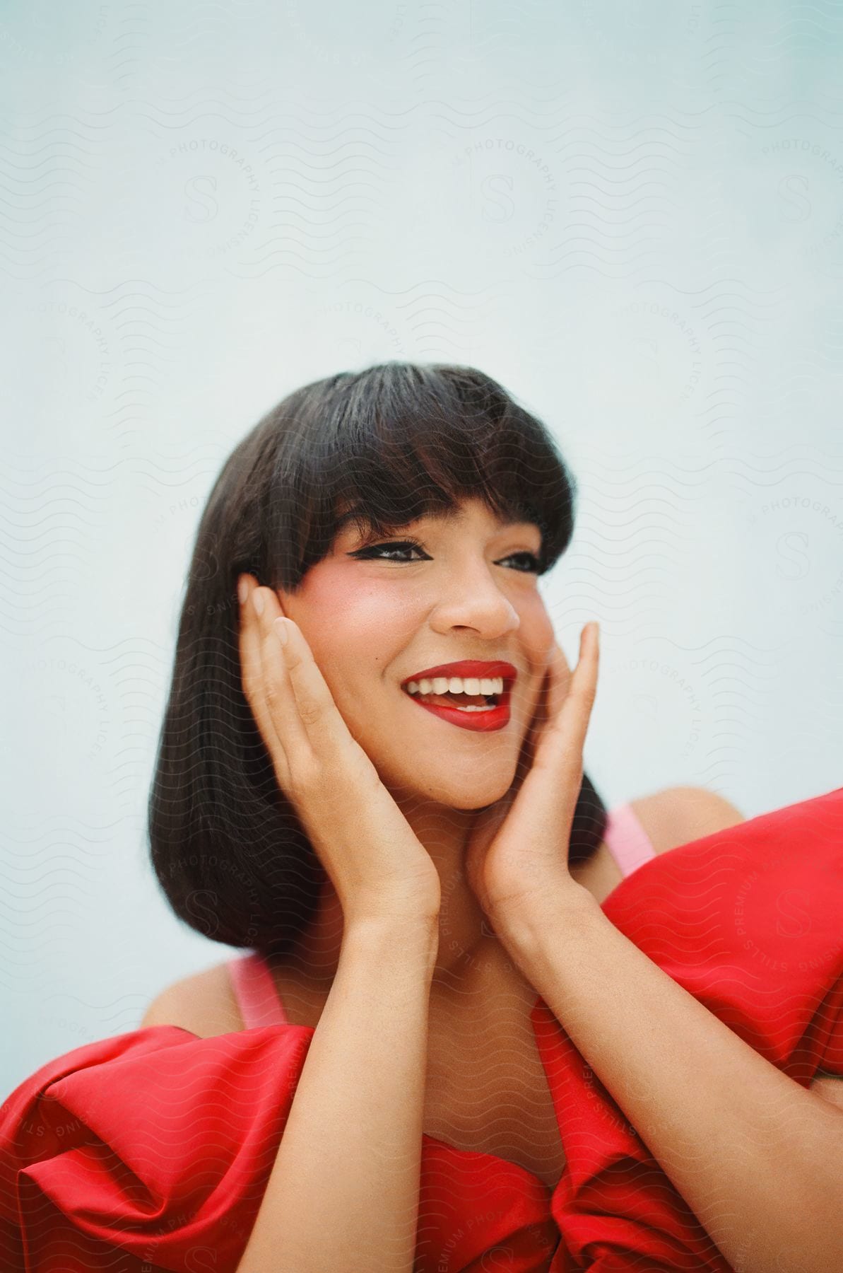 A woman wearing a red party dress and red lipstick smiles with her hands on the sides of her face