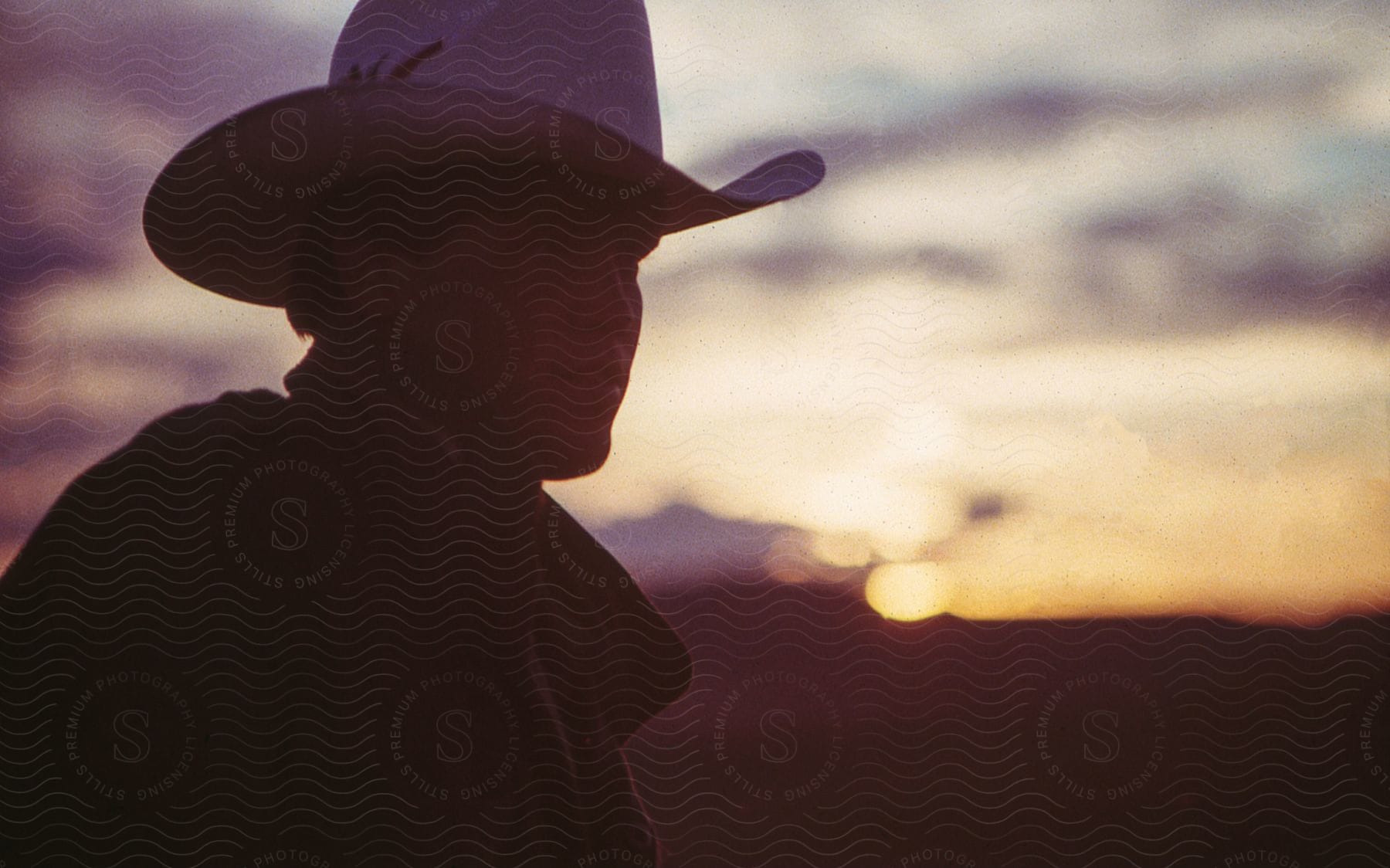 A man wearing a cowboy hat looks to his side as the sunset glows in the distance