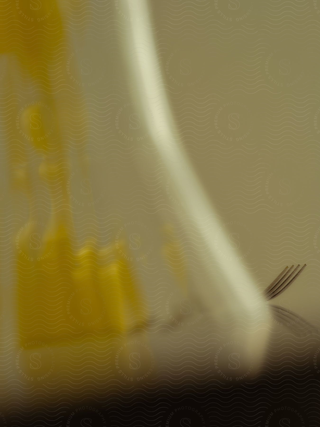 Looking through a gently blurred glass vase, one can observe the bending of light caused by a yellow object. On the right side, a silver fork is mirrored in the reflective surface.