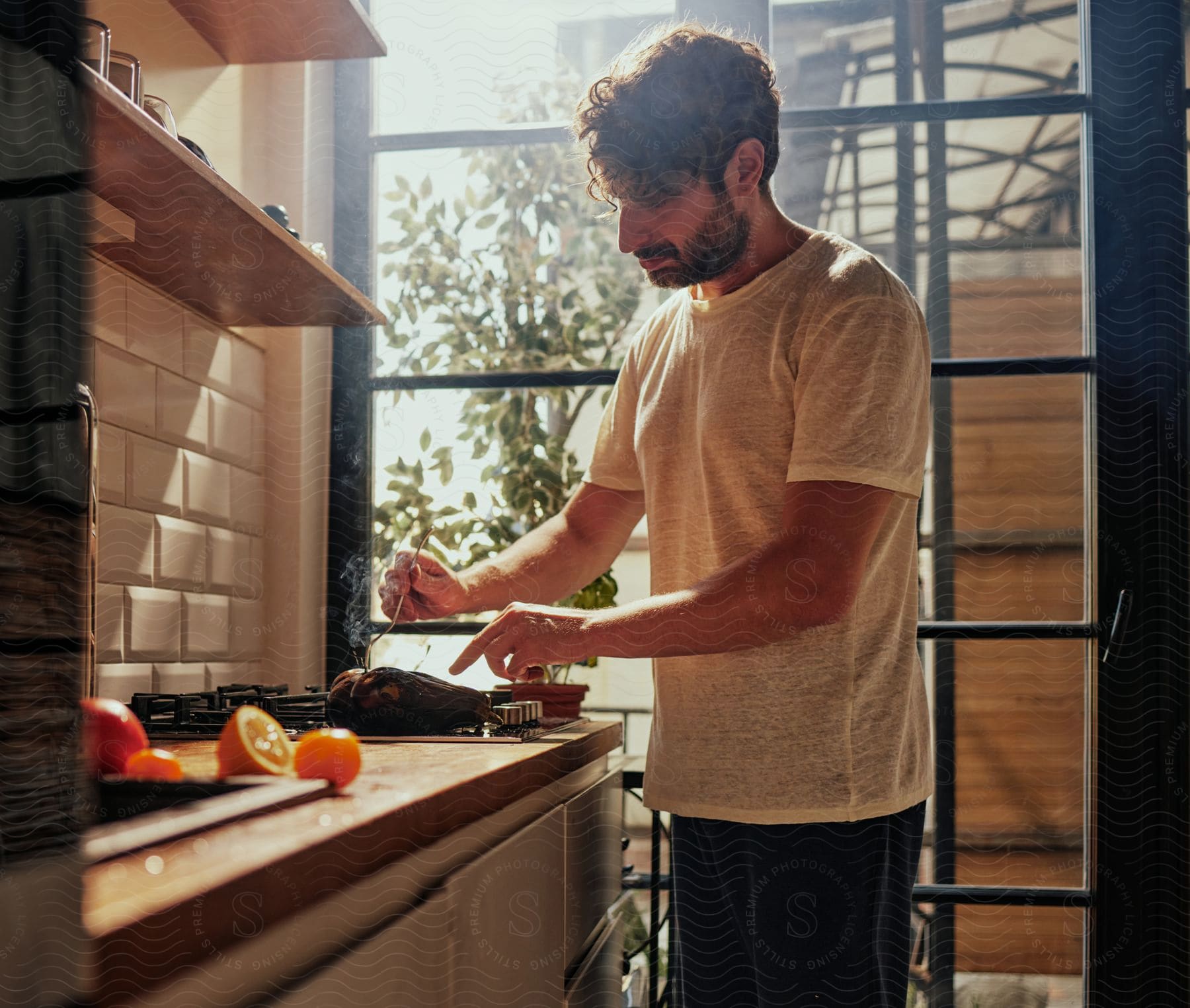 A bearded man, dressed in a white t-shirt and black pants, is cooking eggplants on the stove in the kitchen.