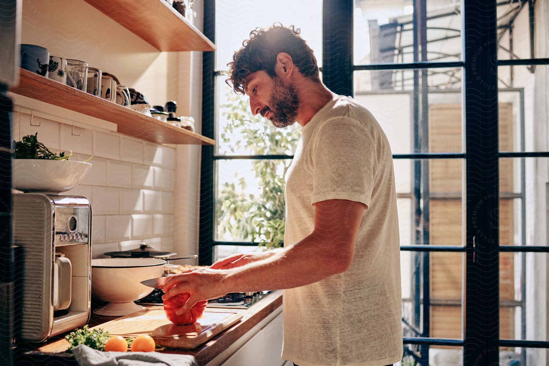 A man in a white shirt prepares food on a kitchen counter with a window view, next to a table of fresh vegetables.