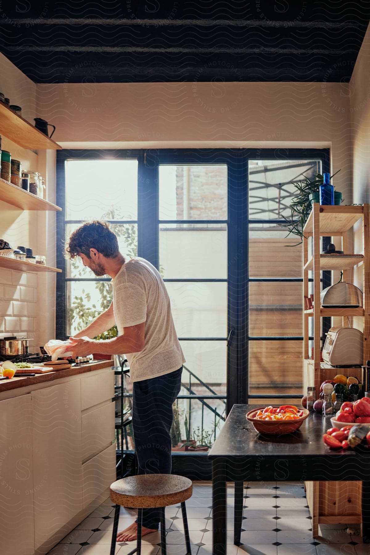 A man in white shirt and black pajamas barefoot is preparing food on a kitchen counter top next to a window, with a table on the right with fresh vegetables.