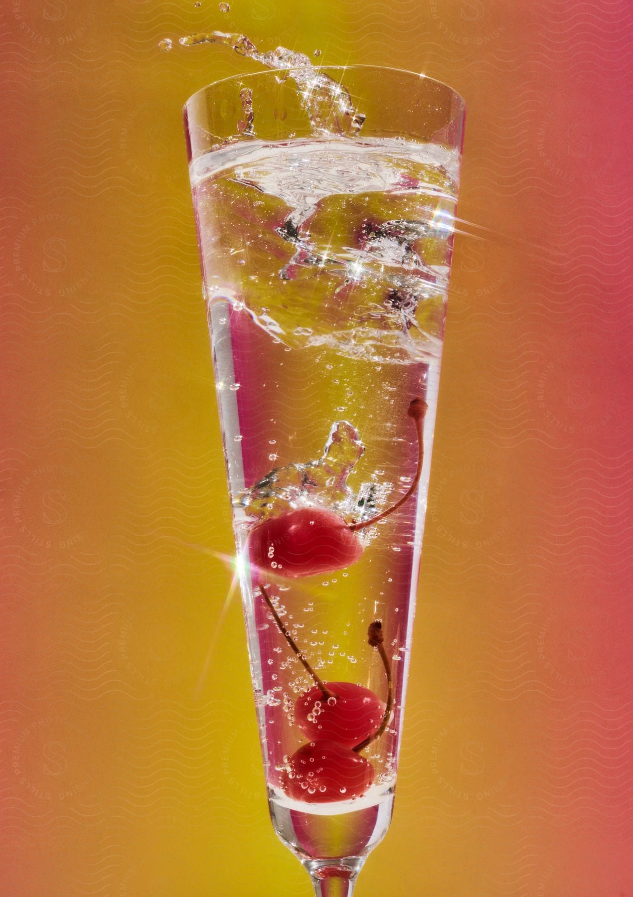 Three cherries with stems dropping into a tall glass of sparkling liquid, creating dynamic splashes, against a gradient yellow to pink background.