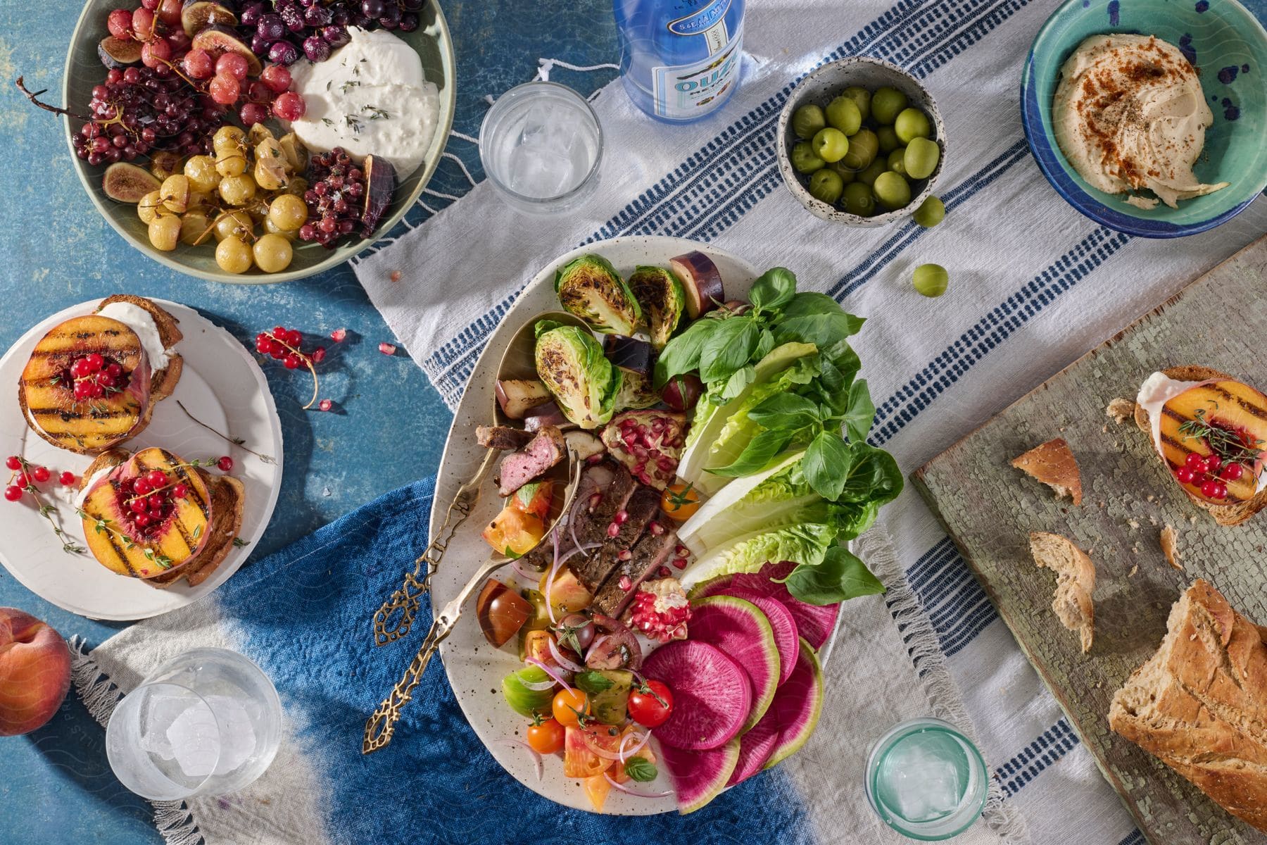 several plates with steak, veggies, fruit, aperitives and desert and three glasses of ice water on a blue table.