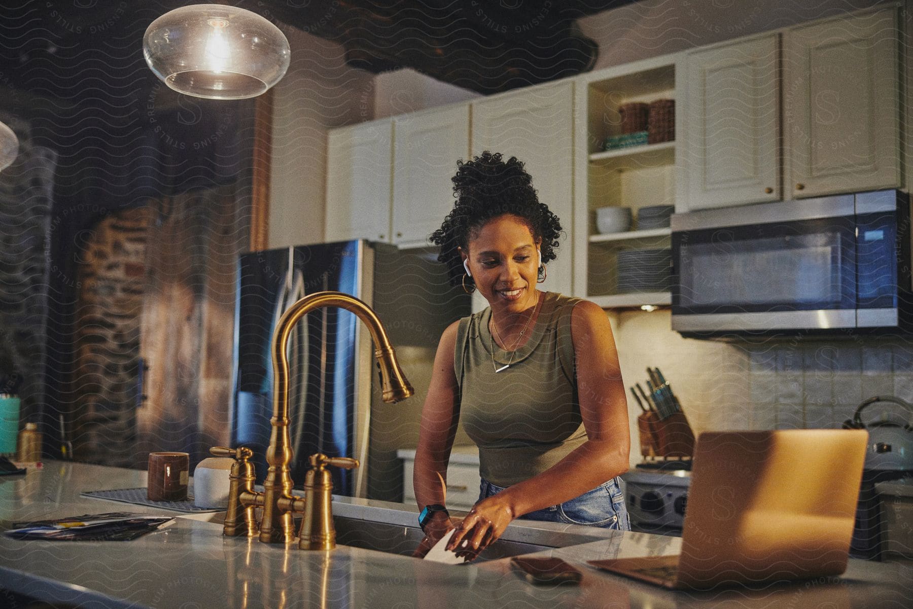 A woman smiling with an airpods while washing dishes in a modern kitchen with white cabinets and looking at the laptop on the counter.