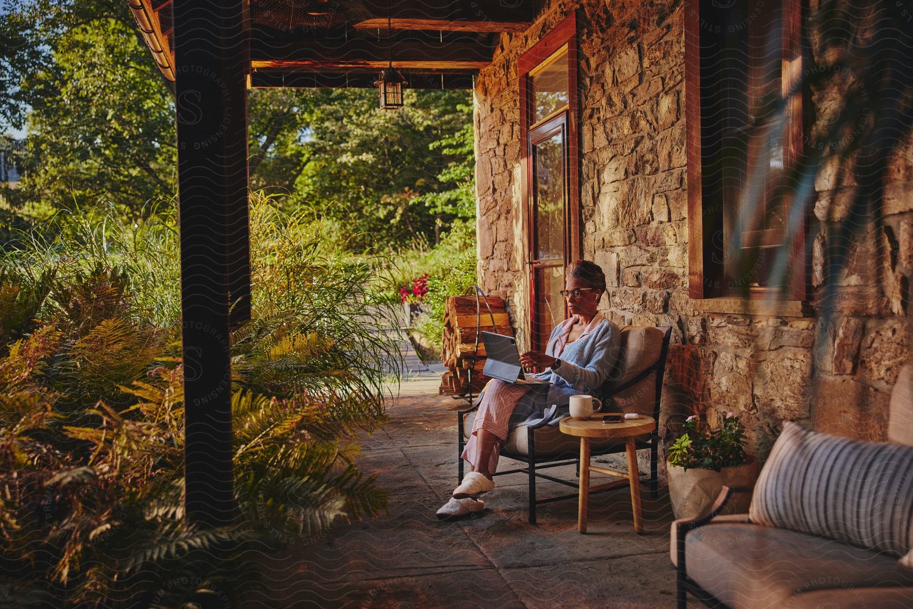 A woman is sitting outside on her patio next to her house in a rural area with a laptop computer