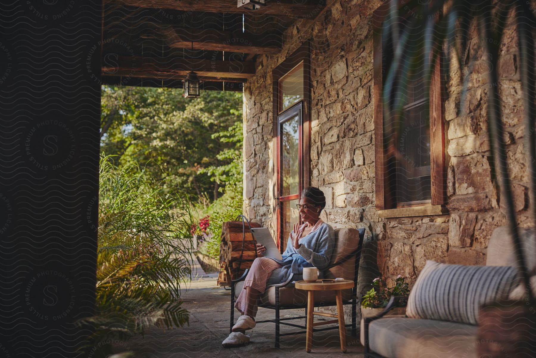 A woman sitting on a chair on a rustic porch, using a tablet, with lush greenery in the background.