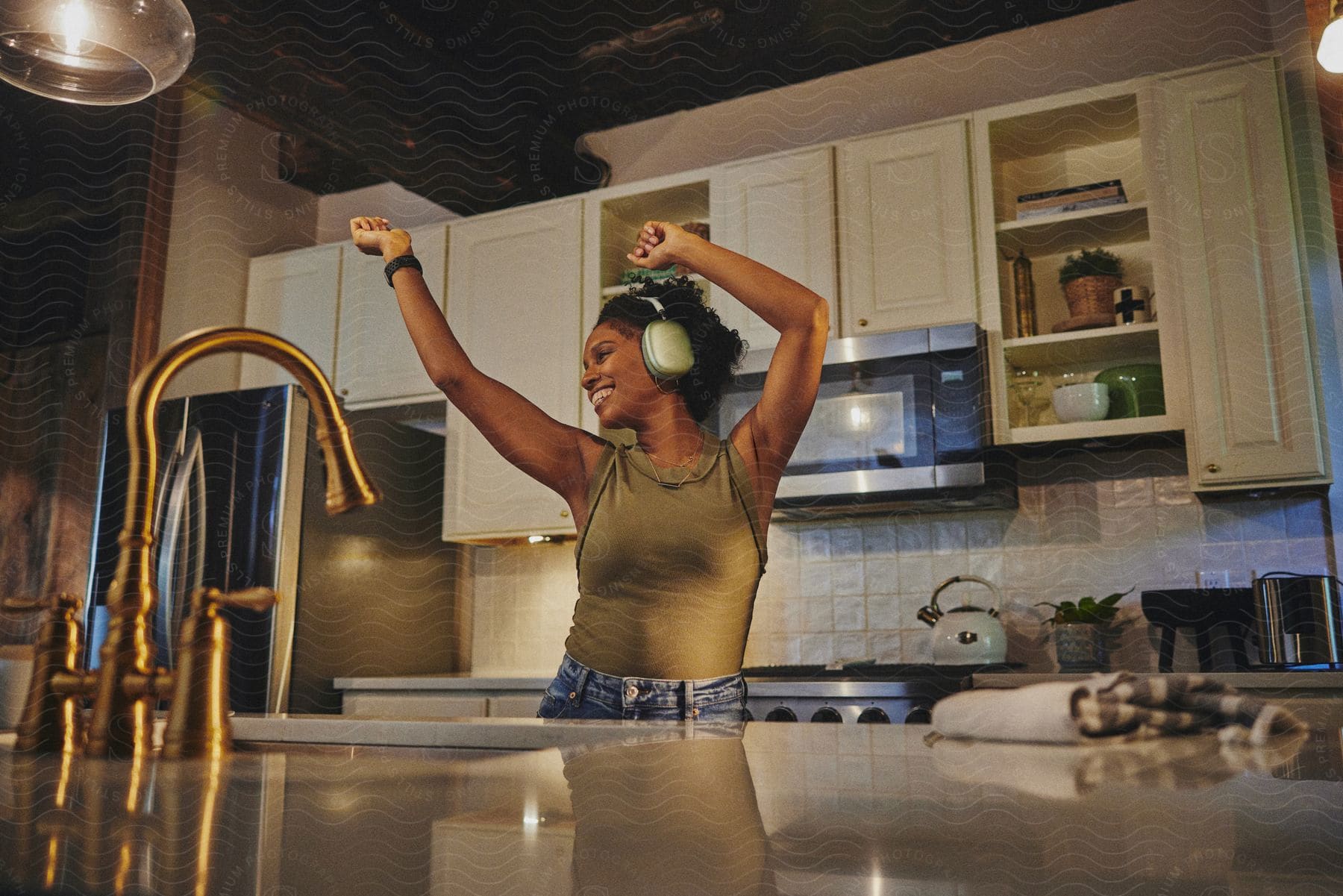 A woman with curly hair, silver headphones, a green top, and jeans dances behind the kitchen island.