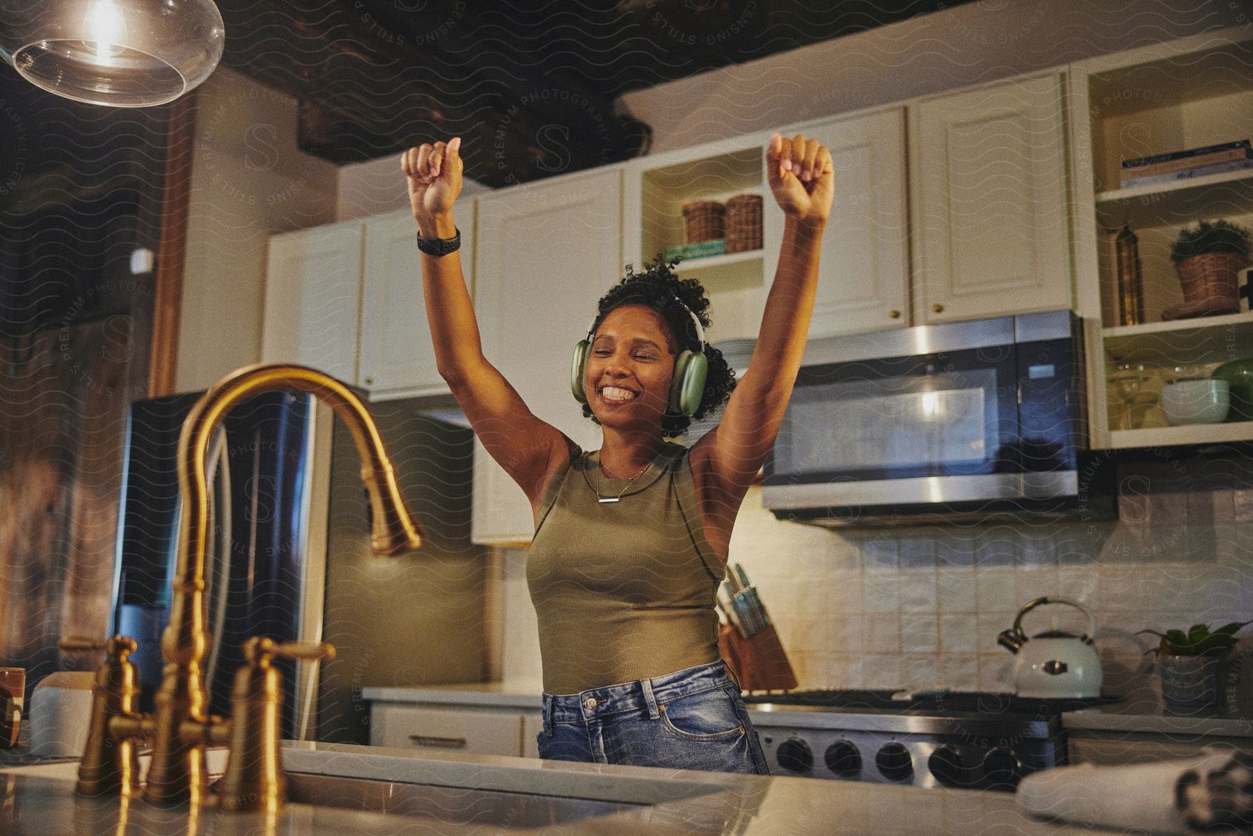 A young woman wearing wireless headphones smiles while dancing to the music in a kitchen.