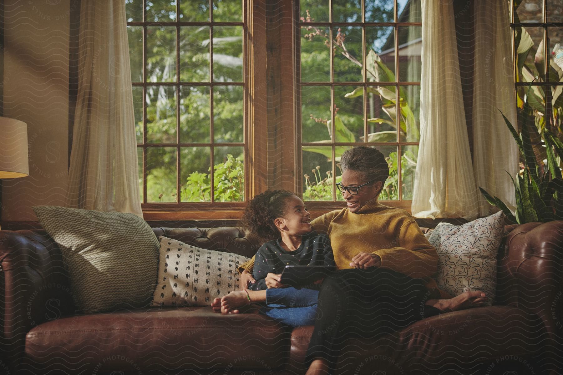 A woman sits on the living room couch laughing with her granddaughter holding a tablet computer