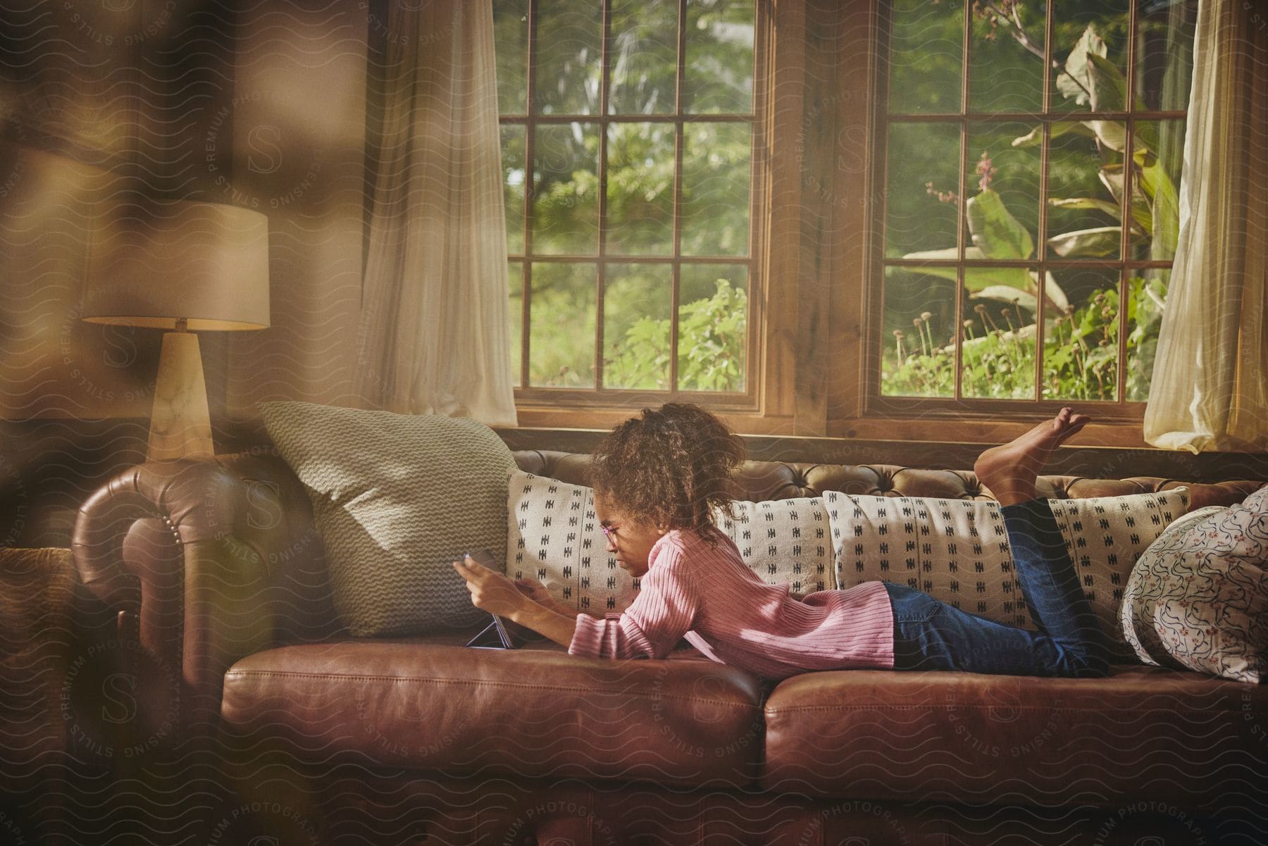 A young girl lies on a couch while holding and staring at a computer tablet.