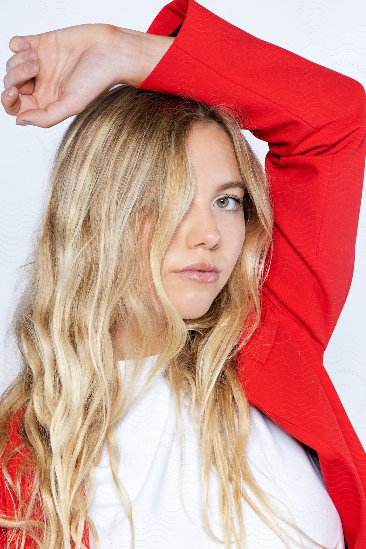 A person with blonde hair wearing a red blazer over a white top, posing with one arm raised and hand behind the head.